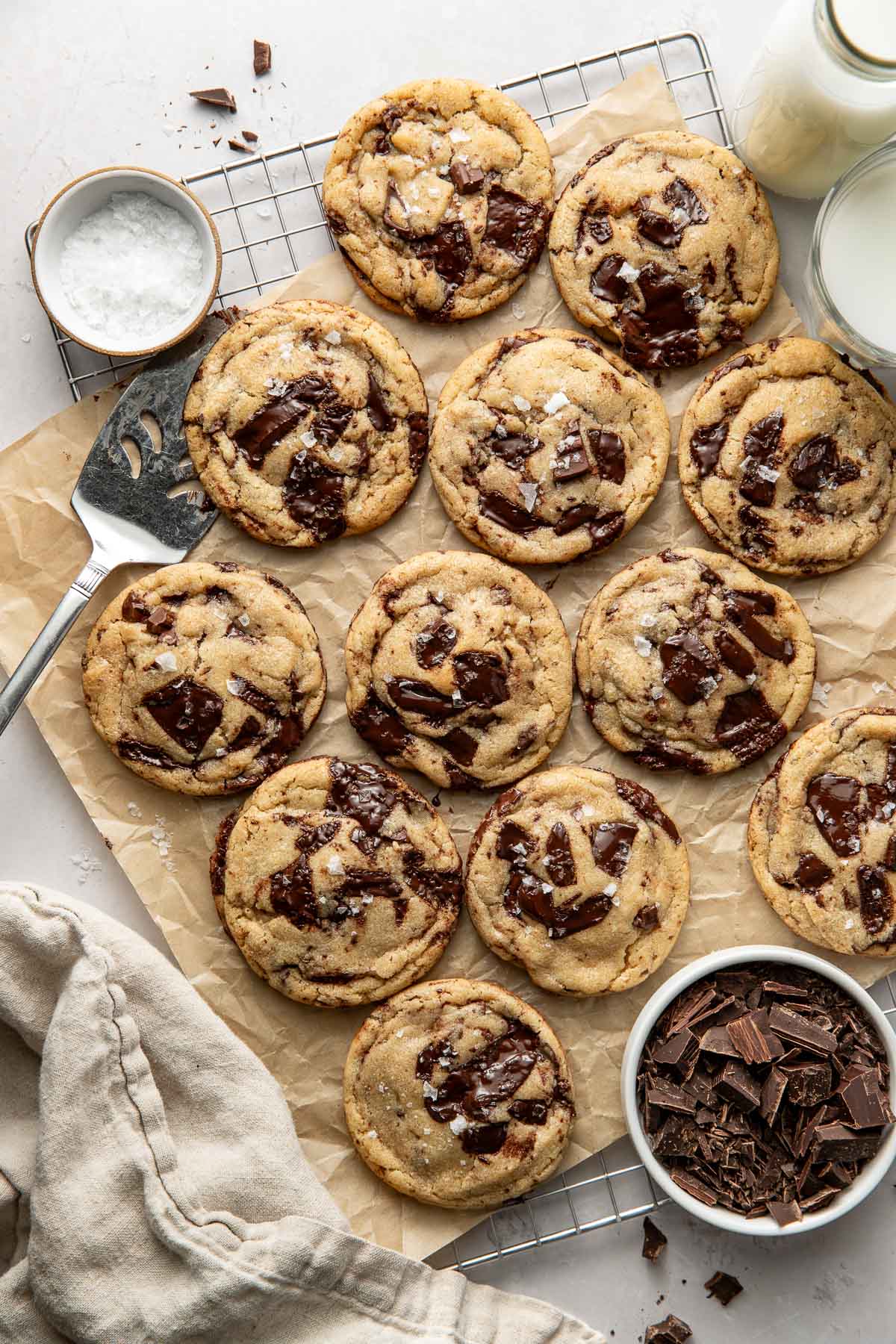 Freshly baked chocolate chip cookies with chunks of chocolate are arranged on parchment paper over a cooling rack, surrounded by a bowl of sea salt, a bowl of chocolate chunks, a spatula, and glasses of milk.