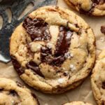 A close-up of gooey chocolate chip cookies topped with flaky sea salt on parchment paper, with a metal spatula and a small bowl of salt visible in the corner.