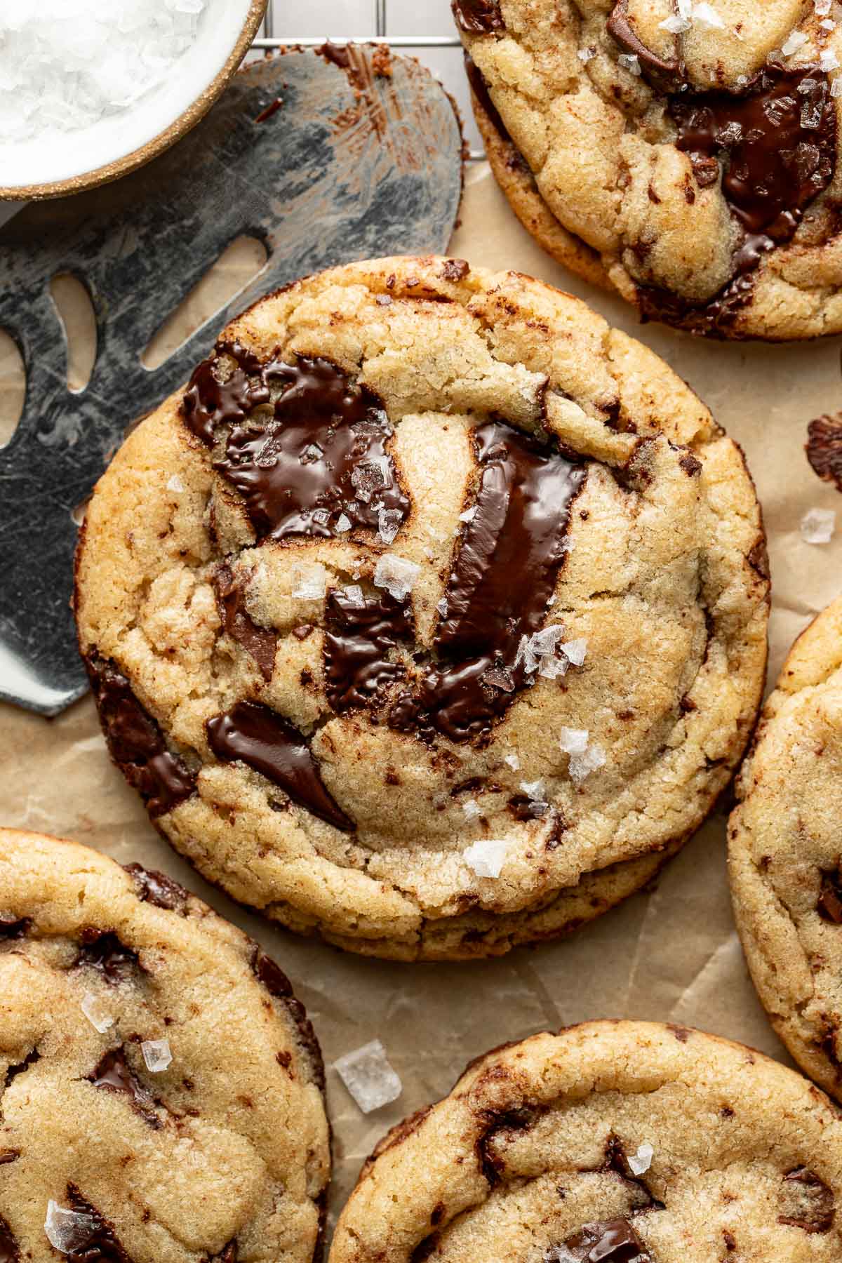 A close-up of gooey chocolate chip cookies topped with flaky sea salt on parchment paper, with a metal spatula and a small bowl of salt visible in the corner.
