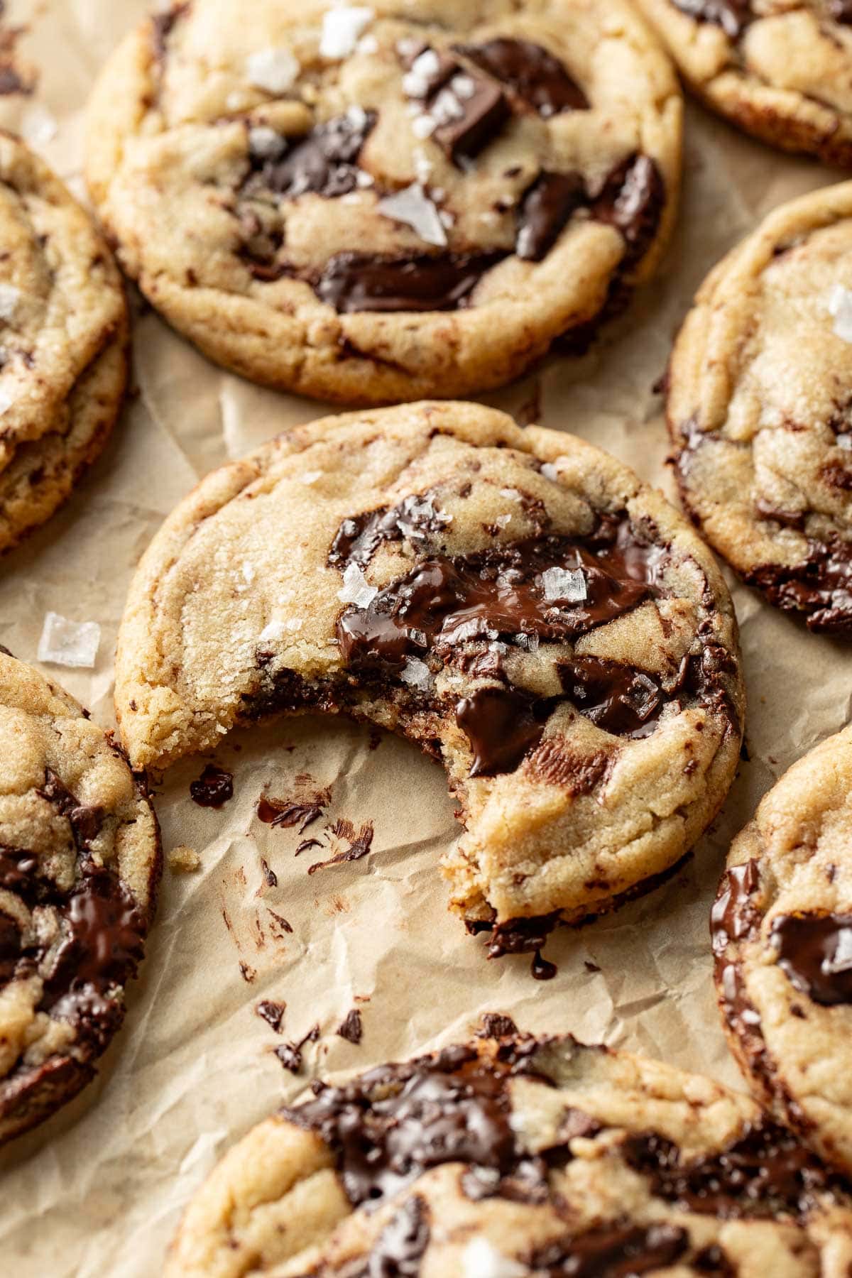 Close-up of chocolate chip cookies on parchment paper, one with a bite taken out. Melted chocolate chunks and flaky sea salt are visible on top of the golden, chewy cookies.