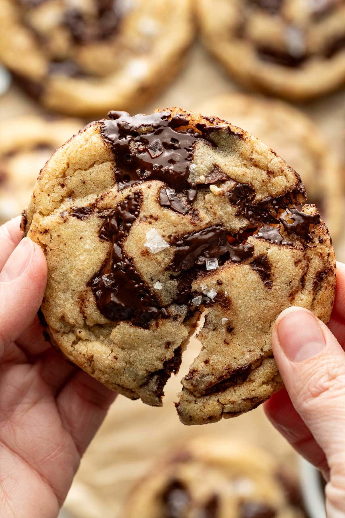 A close-up of hands breaking a chocolate chip cookie in half, revealing gooey melted chocolate and sprinkled sea salt on top. Other cookies are blurred in the background.