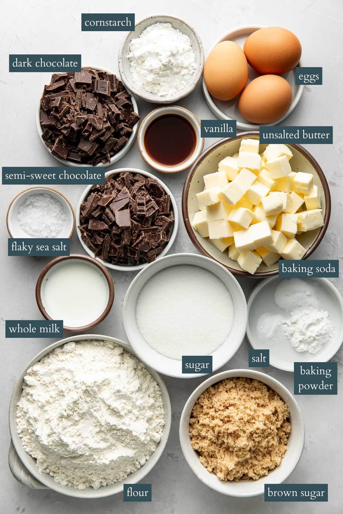 A top view of bowls containing baking ingredients: flour, brown sugar, sugar, whole milk, flaky sea salt, semi-sweet chocolate, dark chocolate, cornstarch, eggs, vanilla, unsalted butter, baking soda, baking powder, and salt.