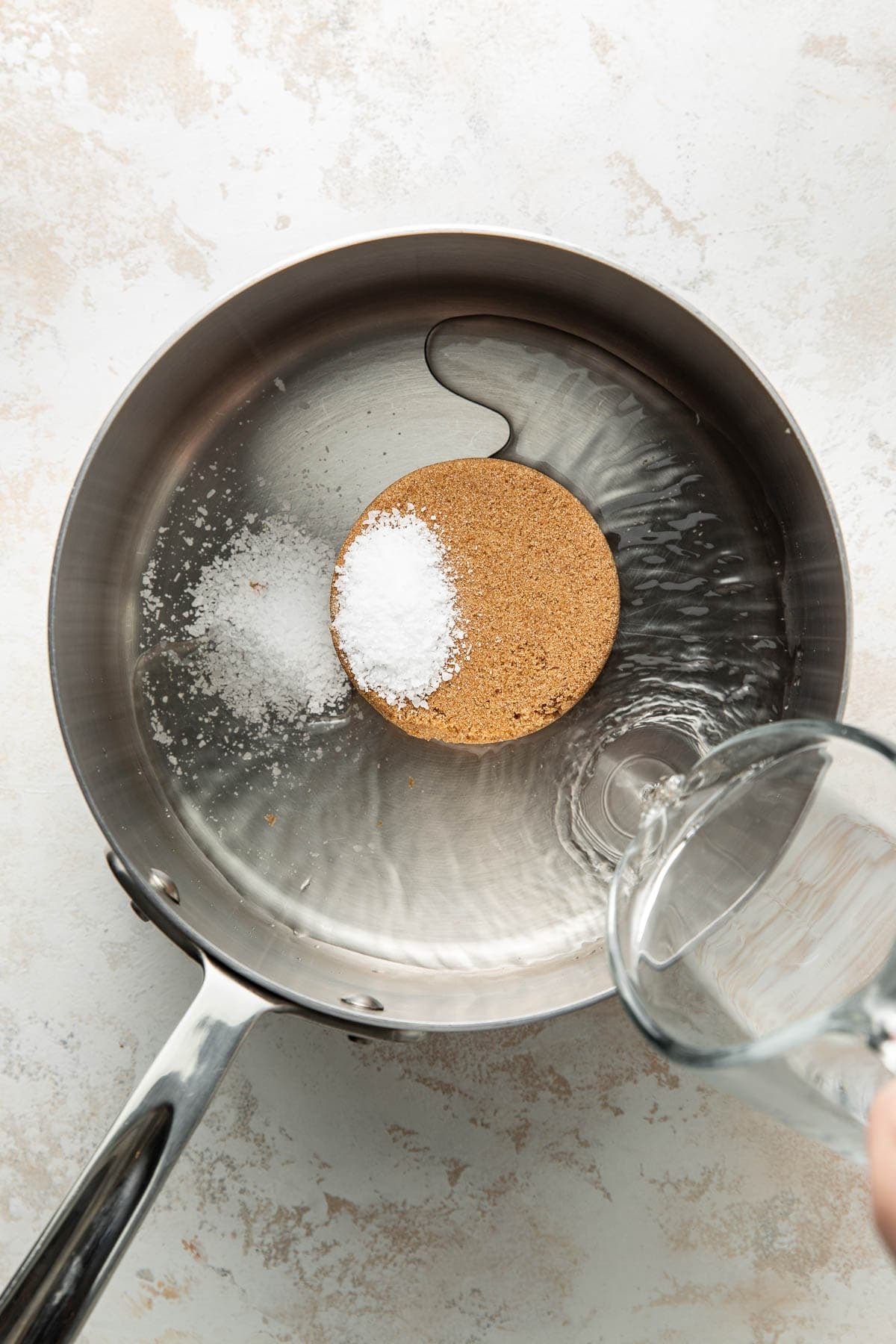 A saucepan with granulated brown sugar, salt, and water inside. A hand is pouring more water into the pan, which is placed on a light-colored surface.