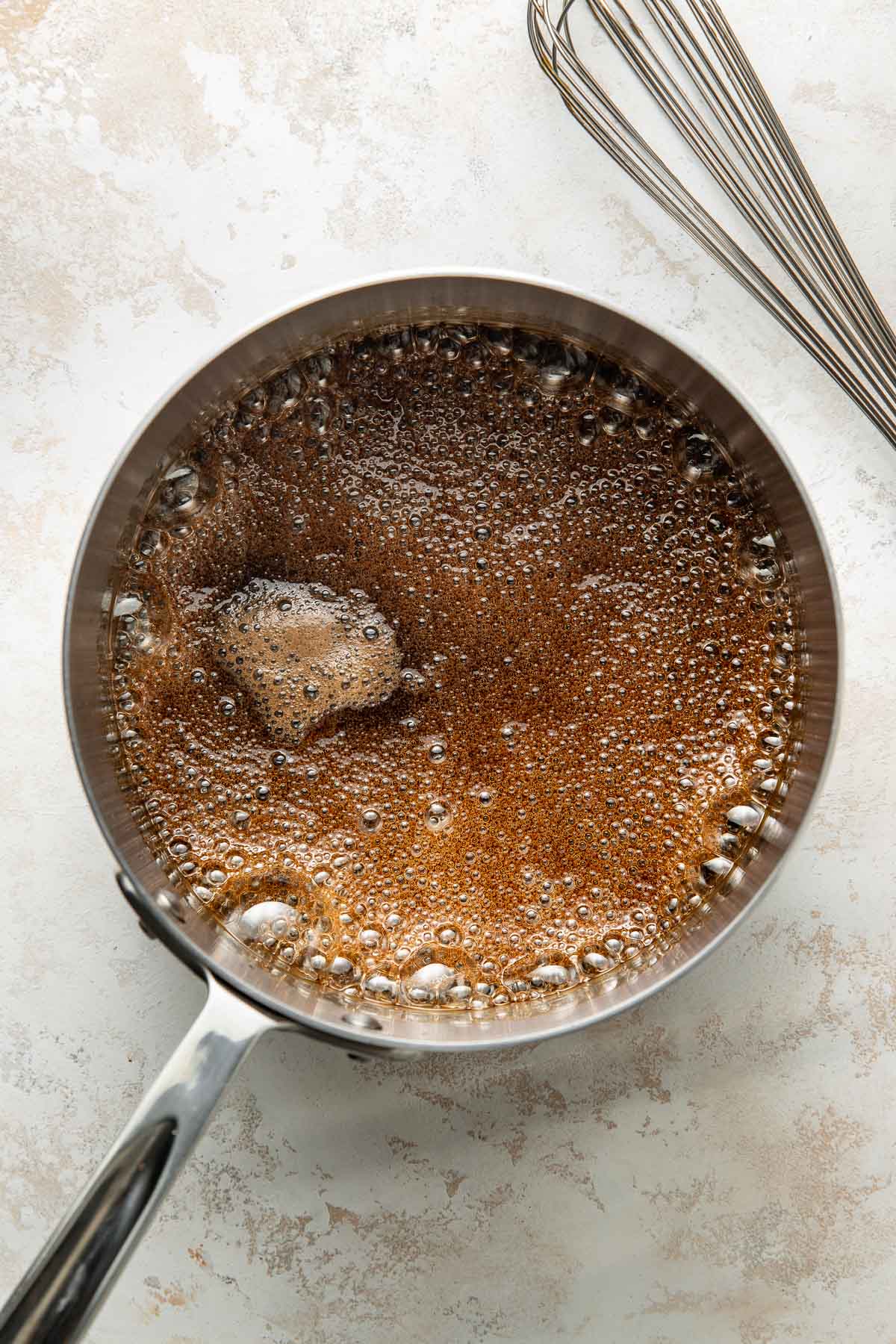 A stainless steel saucepan filled with bubbling brown butter sits on a light countertop next to a metal whisk.