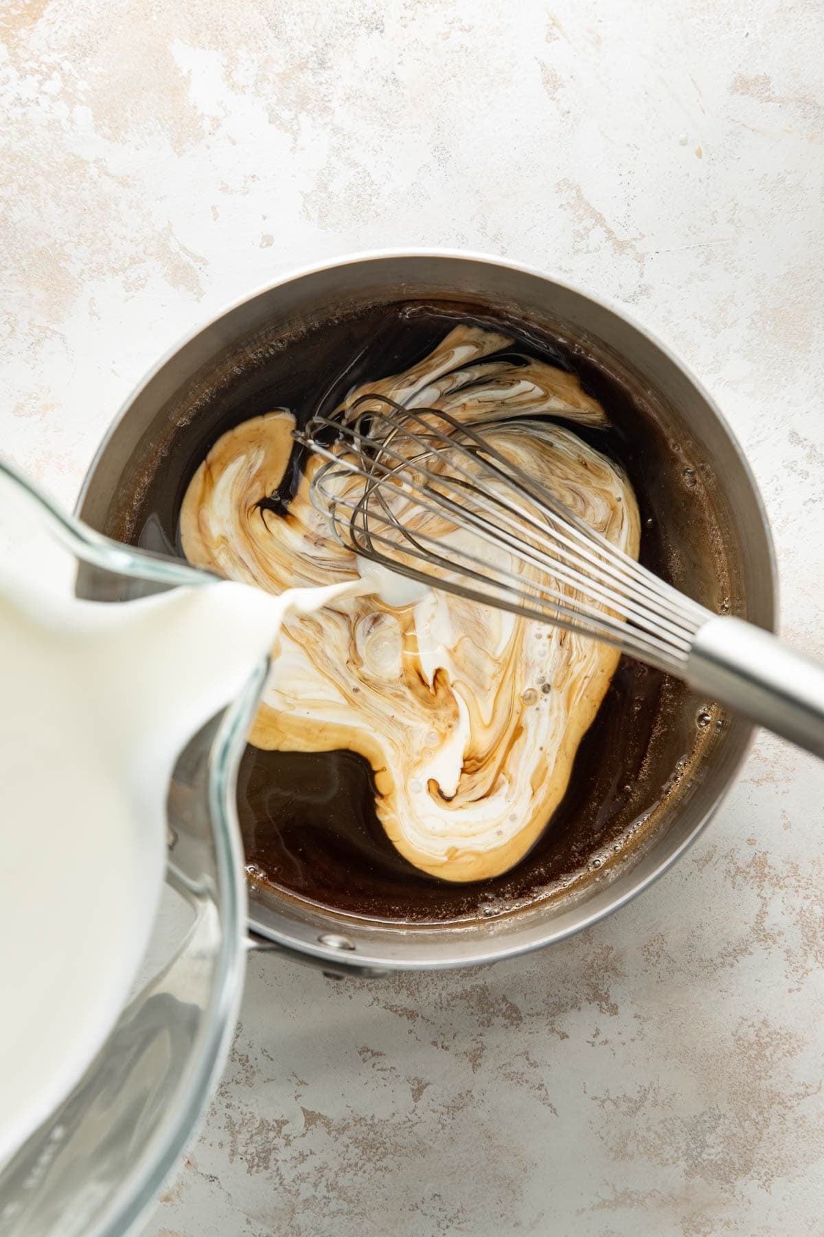 A whisk stirs a mixture of dark liquid and cream in a metal bowl, creating swirling patterns. A glass pitcher pours more cream into the bowl on a light, textured surface.