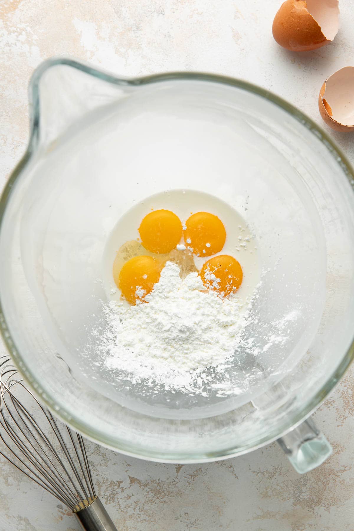 A glass mixing bowl containing four cracked eggs and flour, with eggshells and a metal whisk beside the bowl on a light-colored surface.