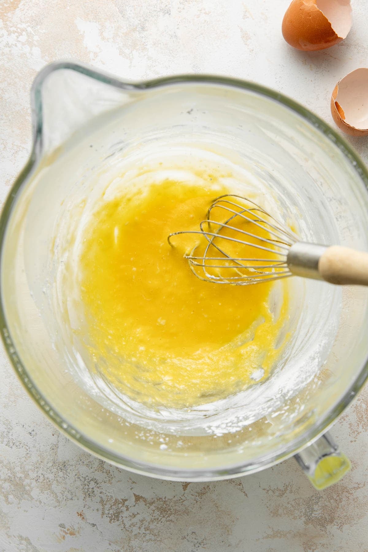 A glass mixing bowl containing a yellow, partially mixed batter and a metal whisk with a wooden handle. Eggshells are visible on the countertop in the upper right corner.