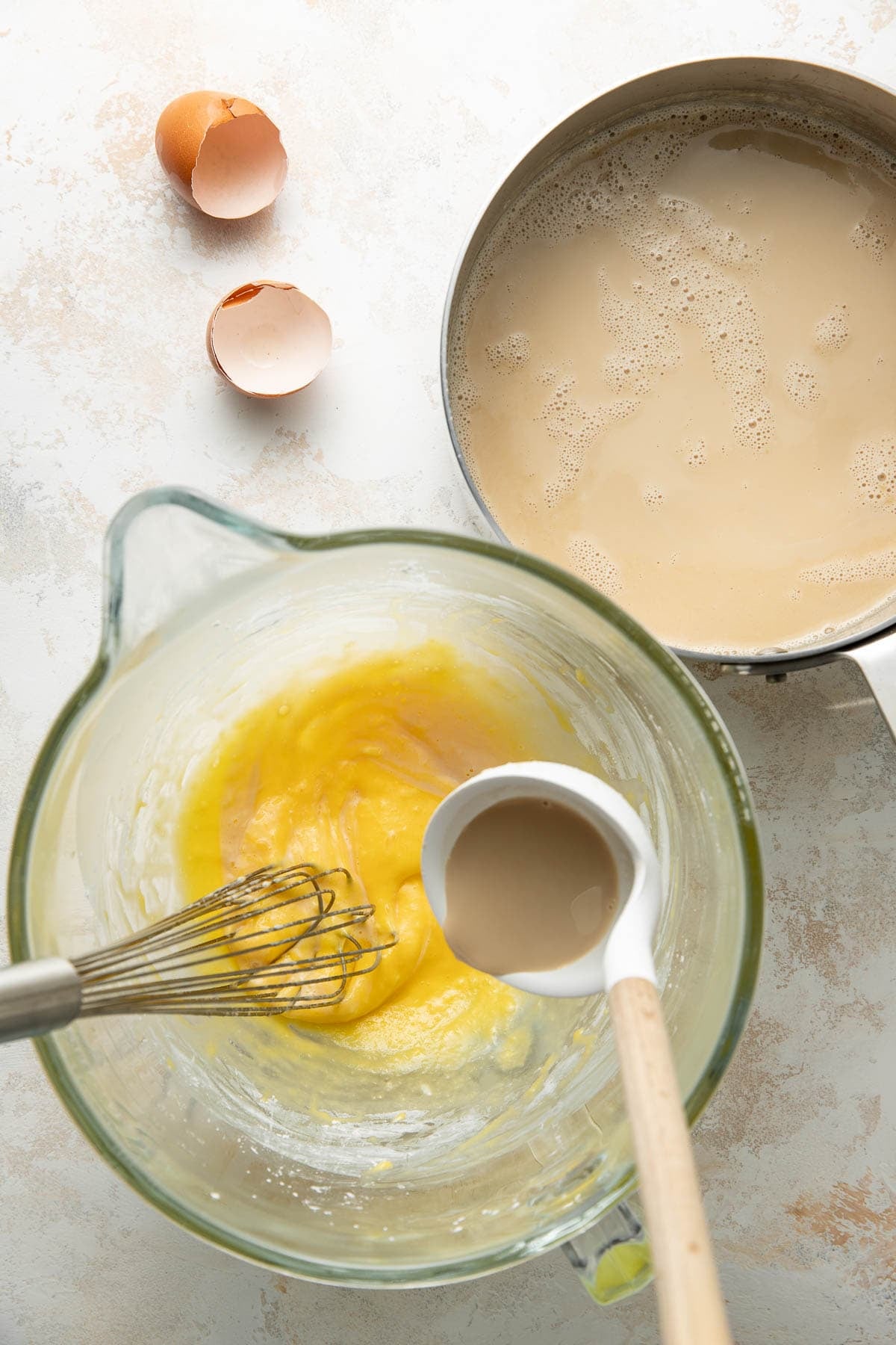 A whisk rests in a glass bowl of beaten eggs and sugar, next to a pot of foamy liquid. A ladle is pouring liquid into the bowl. Two cracked eggshells are on the countertop.
