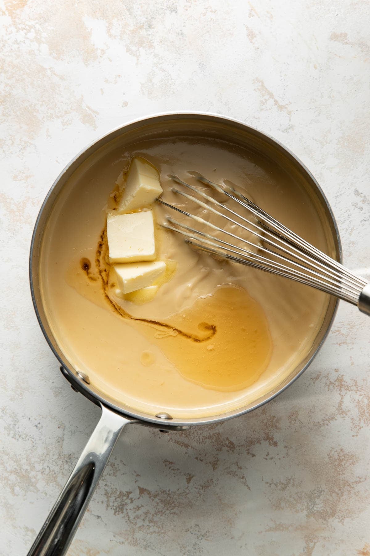 A saucepan filled with creamy sauce, a few pieces of butter, and a splash of vanilla extract being whisked together on a light-colored surface.