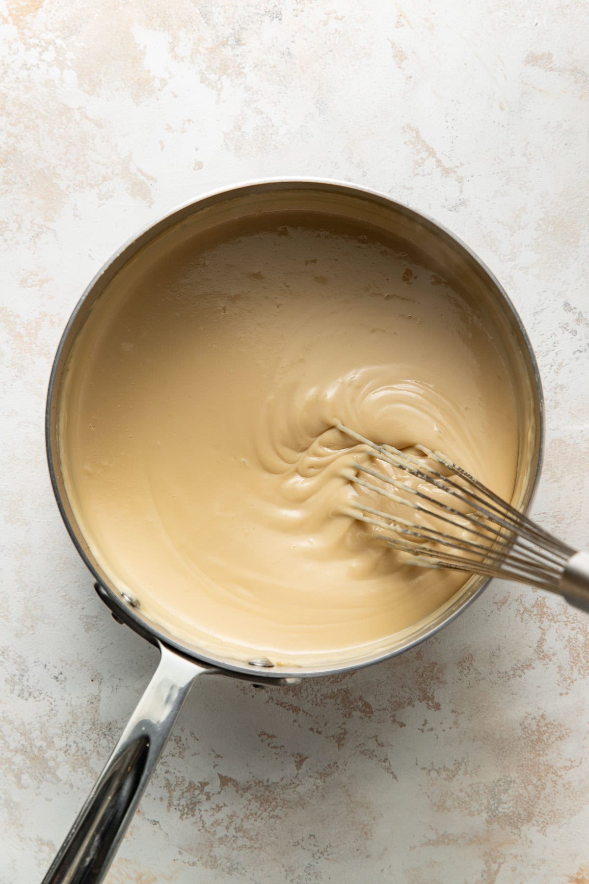 A metal saucepan filled with creamy beige sauce being whisked with a metal whisk, on a light-colored countertop.