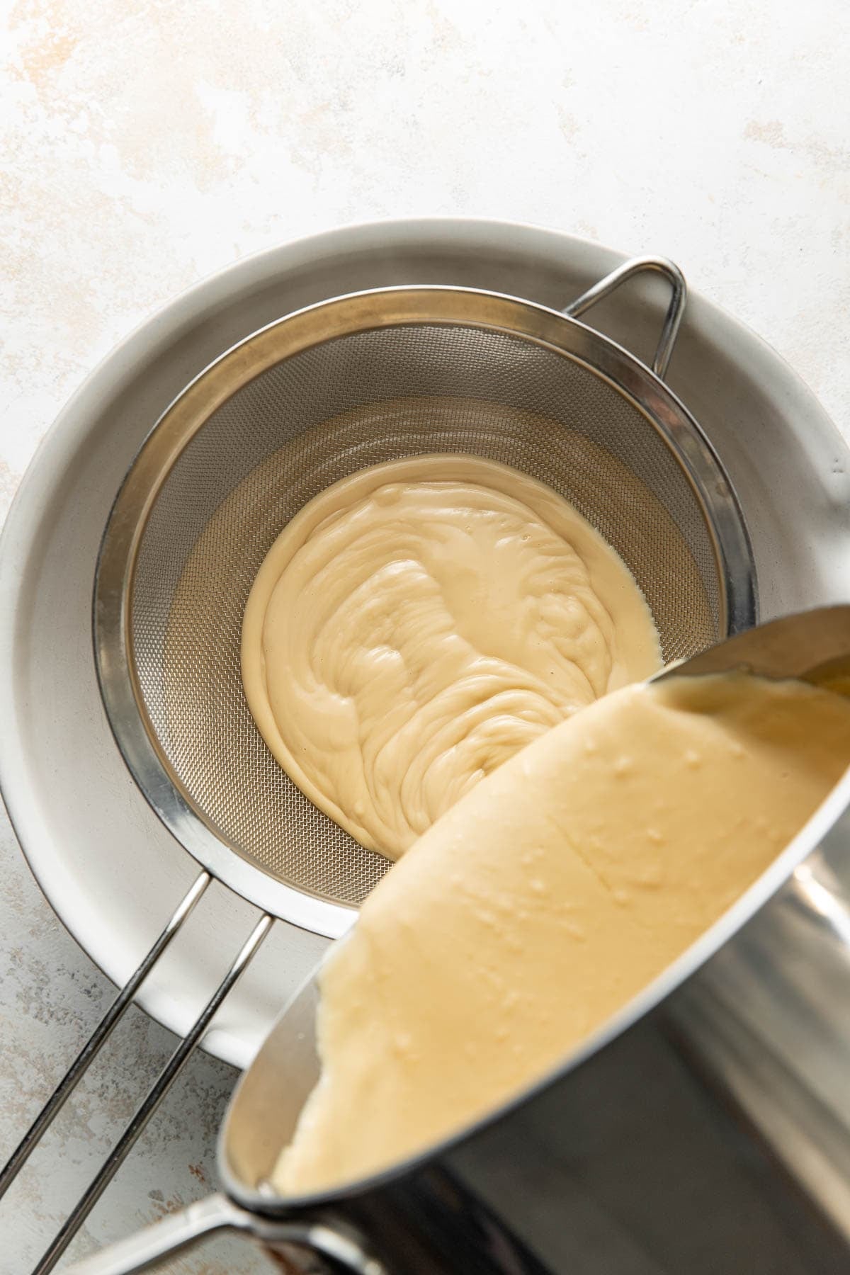 A metal pot pours creamy batter through a fine mesh strainer set over a large white bowl, filtering the mixture as it flows.
