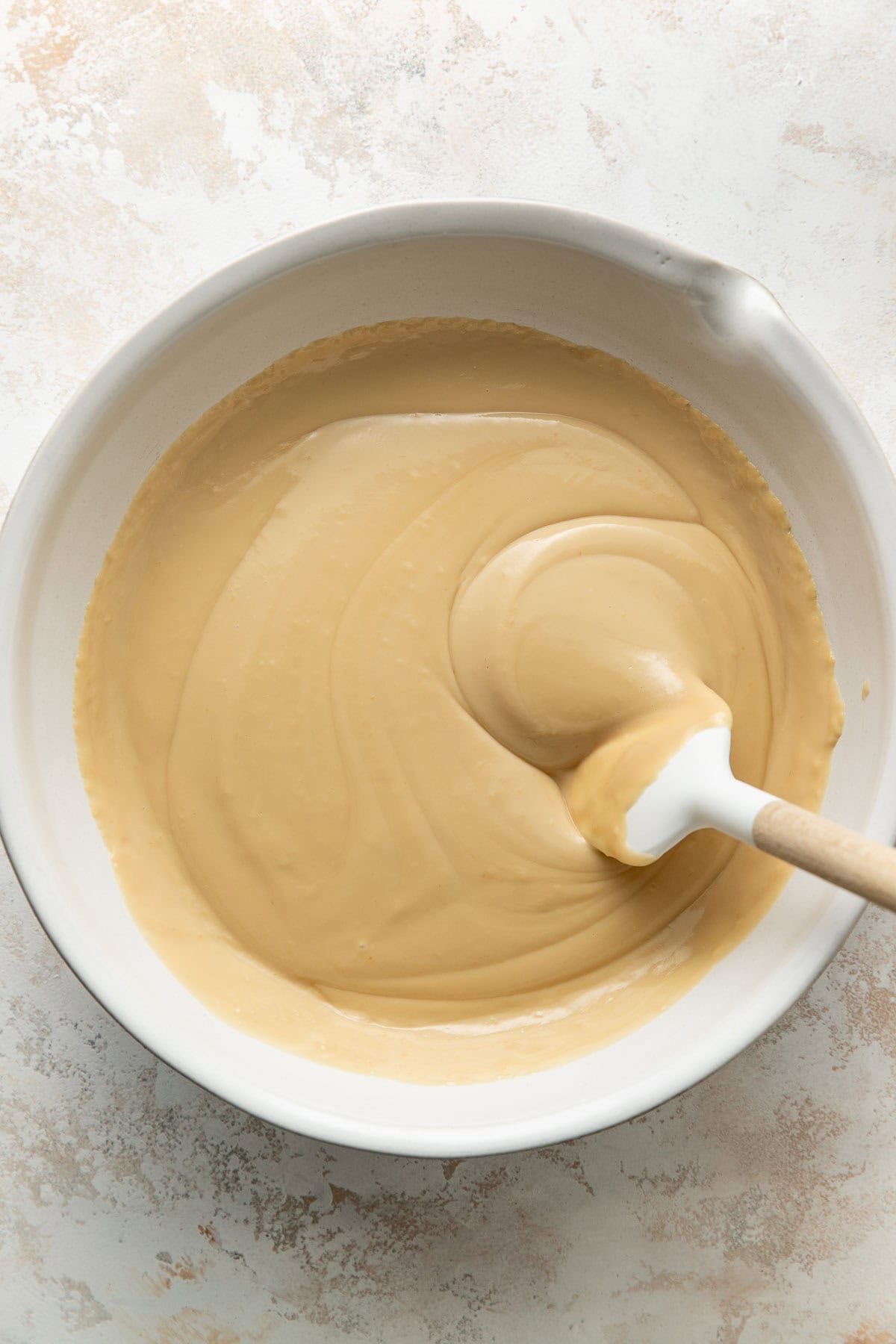 A mixing bowl filled with creamy, light brown batter, with a white spatula stirring the mixture. The bowl is placed on a light, textured surface.