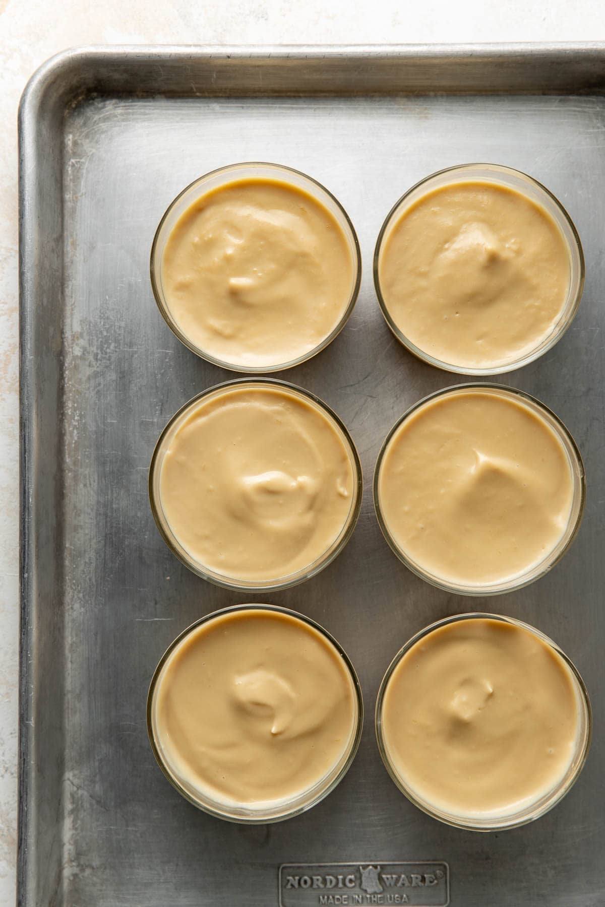 Six glass ramekins filled with smooth, light brown custard are arranged in two rows on a metal baking sheet. The surface of each custard is glossy and slightly uneven.