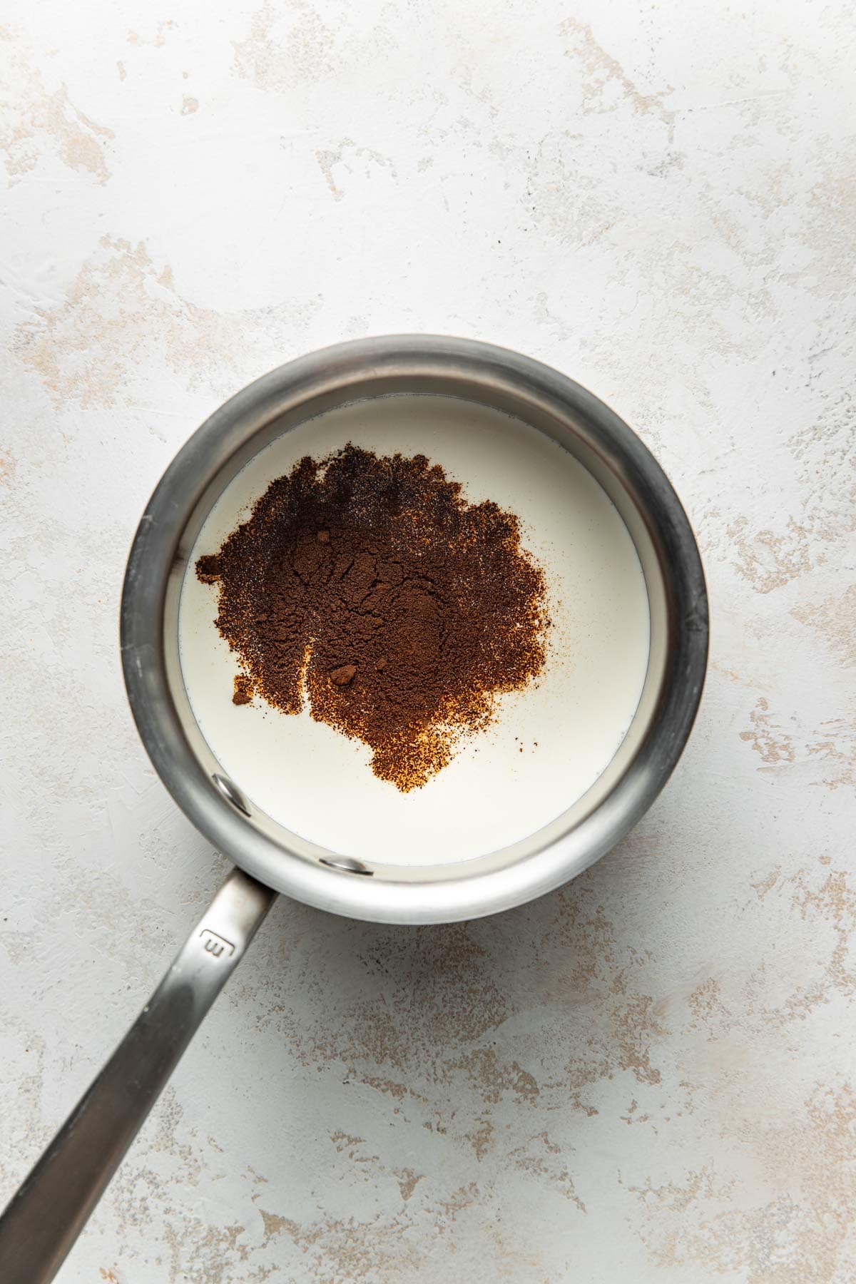 A stainless steel saucepan filled with cream and a pile of ground coffee sits on a light, textured surface.