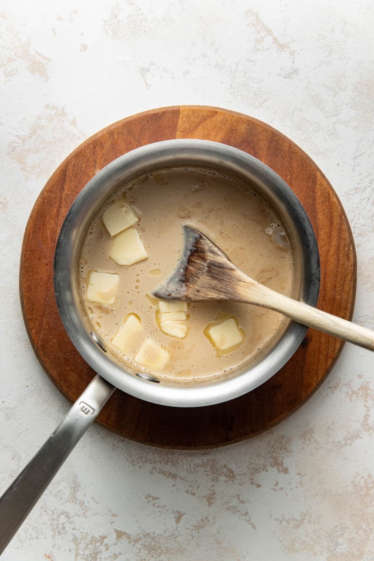 A saucepan with a creamy liquid and several butter slices, being stirred with a wooden spoon, sits on a round wooden board atop a light textured surface.