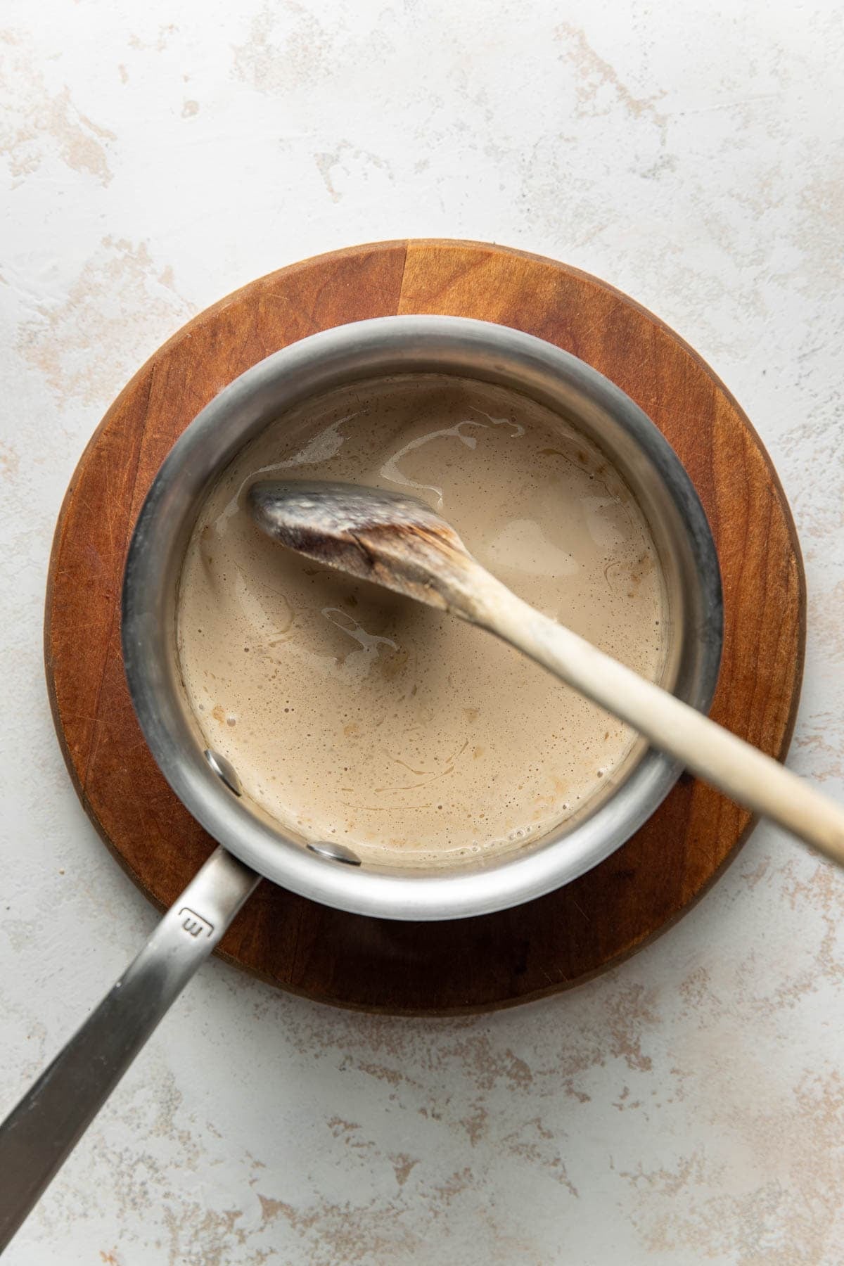 A saucepan filled with a light brown creamy liquid sits on a wooden board, with a wooden spoon resting inside and across the pot.
