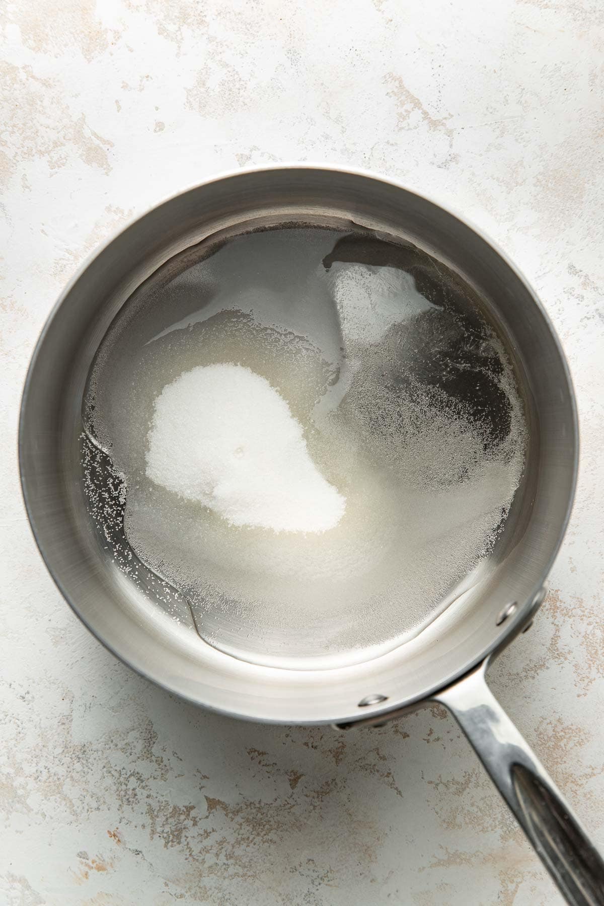 A stainless steel saucepan contains water and a mound of granulated white sugar, partially dissolved, on a light textured surface.