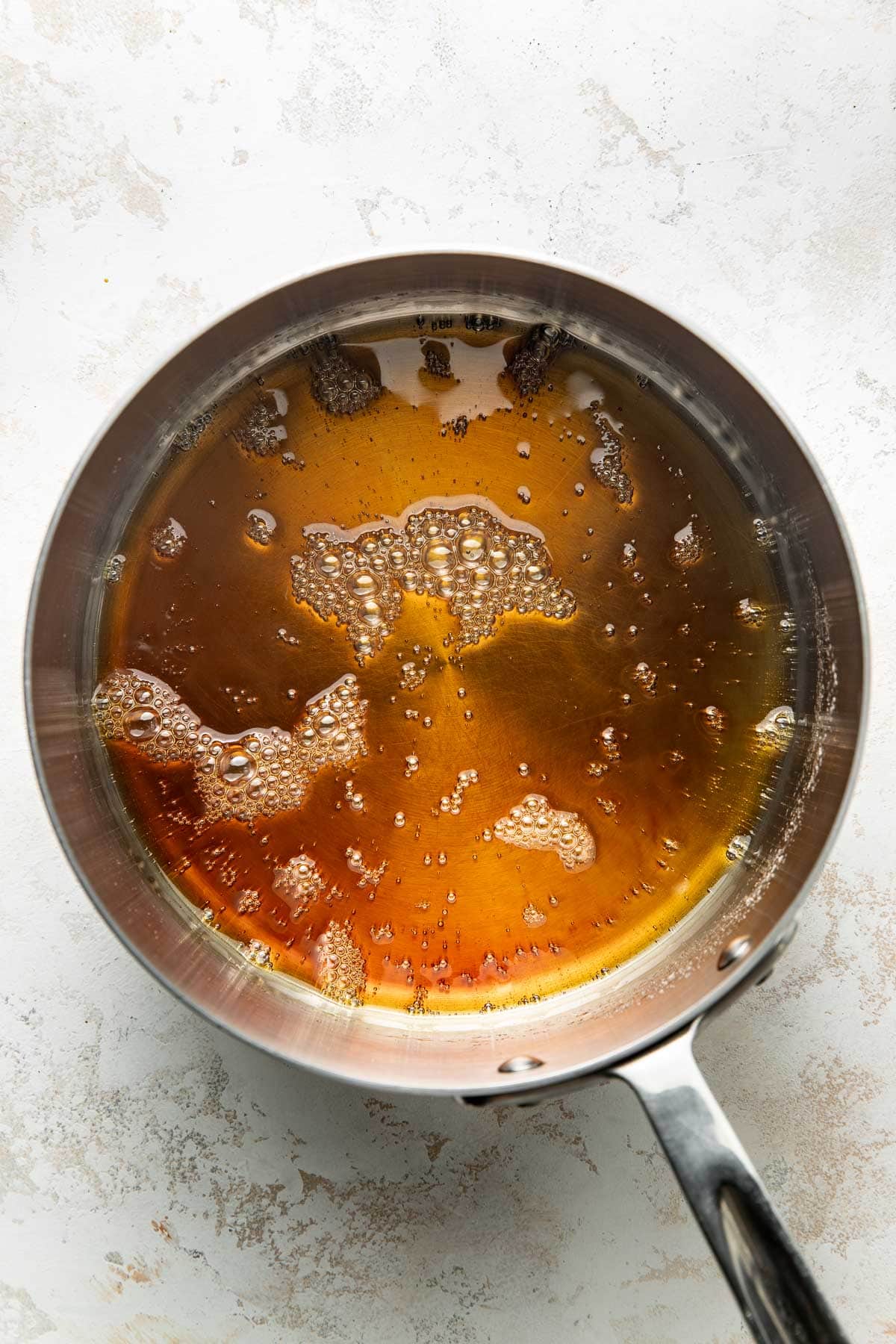 A stainless steel saucepan filled with golden brown melted butter, showing bubbles on the surface, sits on a light, textured countertop.