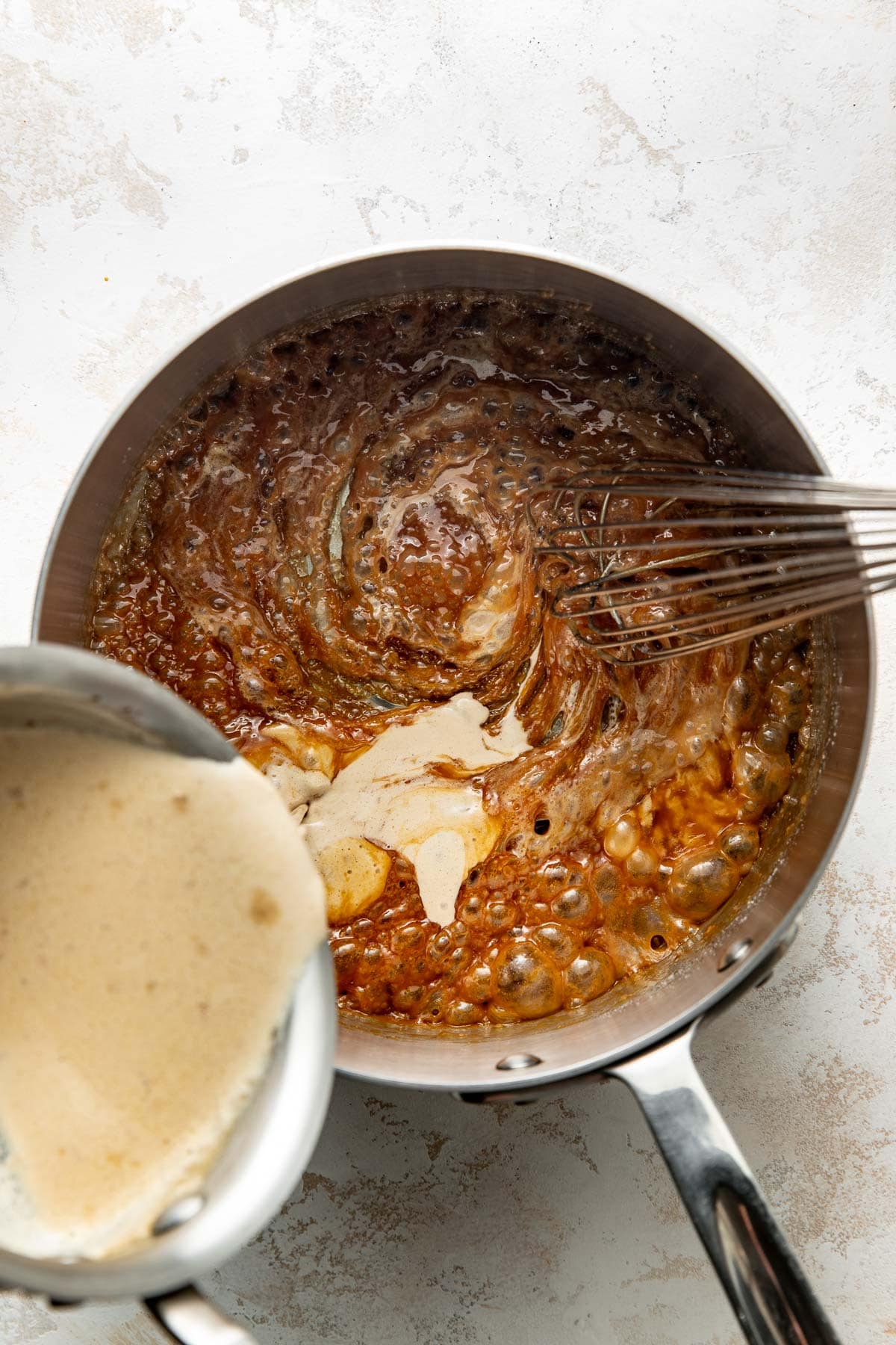A saucepan with bubbling brown mixture being whisked, while a creamy liquid is being poured in from a small pot, on a white surface.