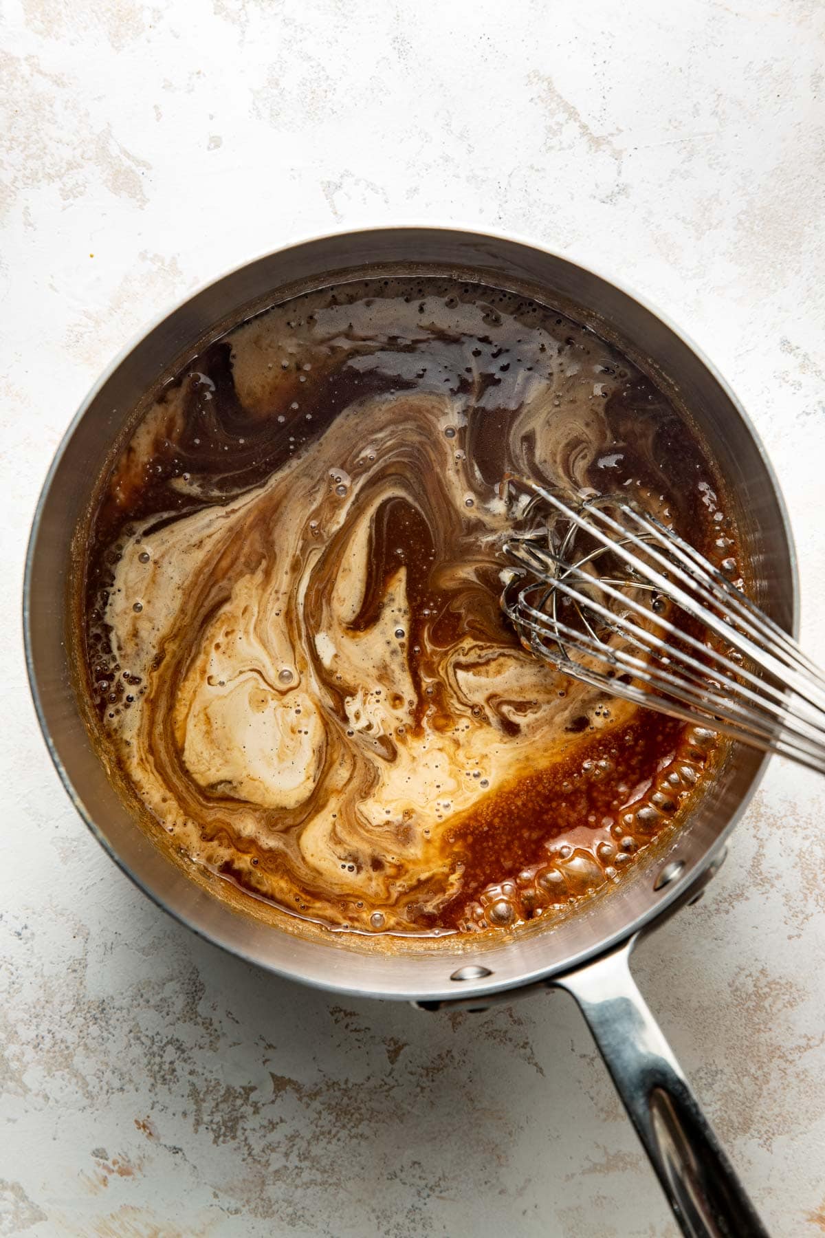 A metal saucepan filled with a swirling mixture of light and dark brown liquid is being whisked with a metal whisk on a light textured surface.