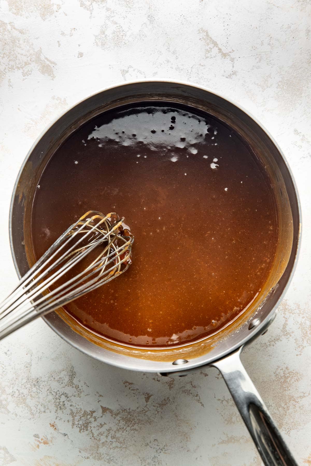 A stainless steel saucepan filled with a smooth, glossy brown sauce being whisked with a metal whisk on a light, textured surface.