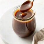A glass jar filled with caramel sauce sits on a white surface, with a spoon held above it dripping caramel back into the jar. A beige cloth is partially visible in the foreground.