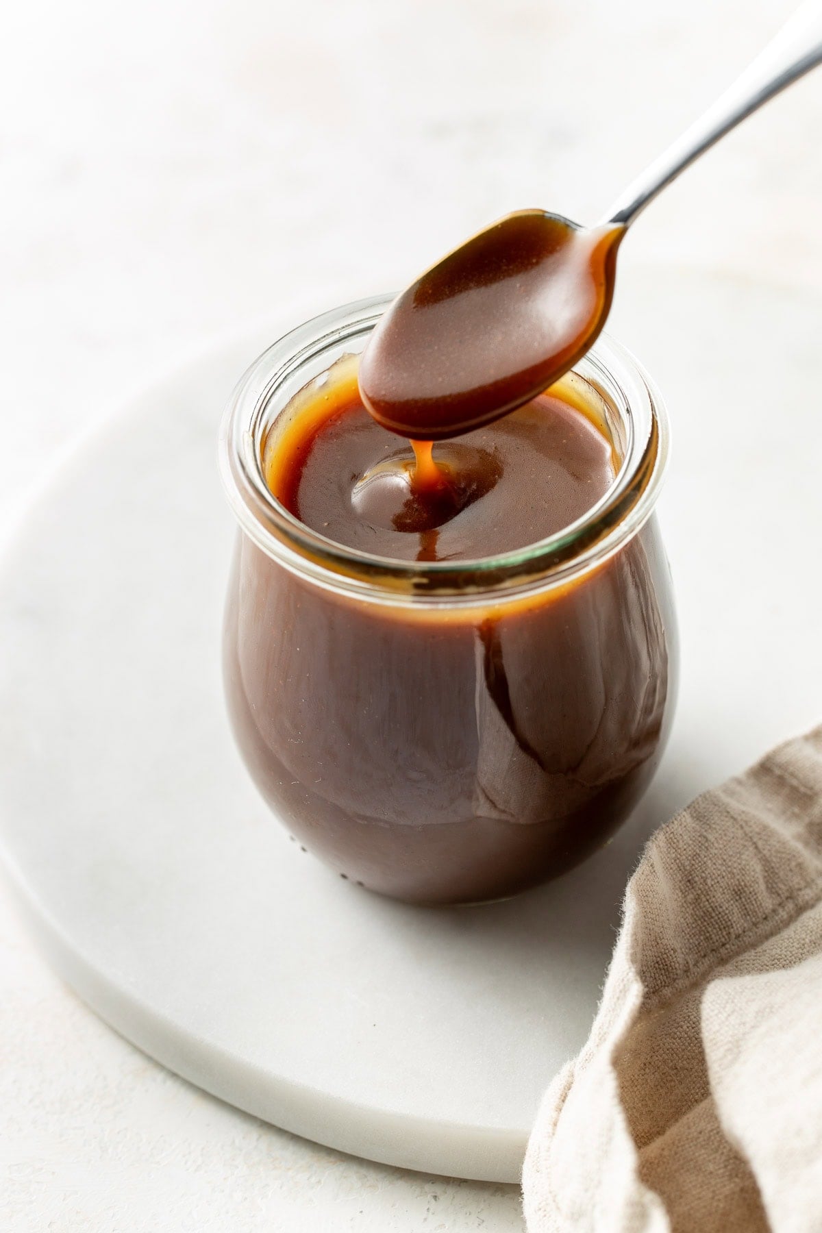 A glass jar filled with caramel sauce sits on a white surface, with a spoon held above it dripping caramel back into the jar. A beige cloth is partially visible in the foreground.