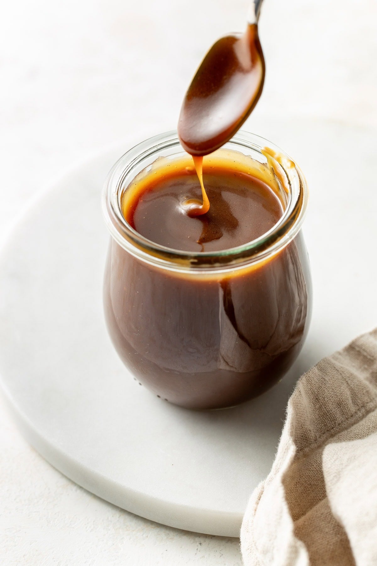 A spoon drizzles caramel sauce into a small glass jar filled with caramel, placed on a white round board next to a beige cloth.