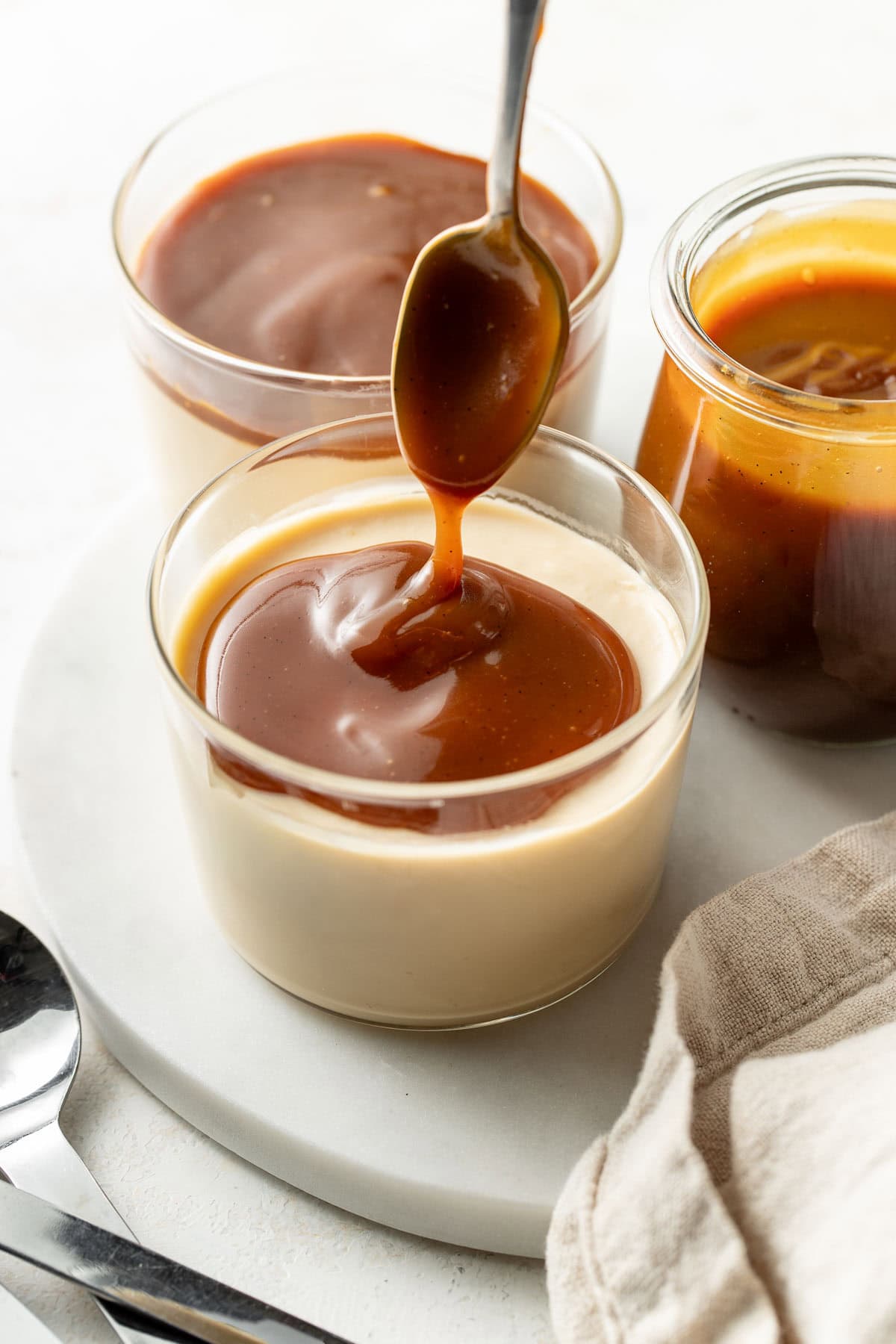 Glass cups filled with creamy panna cotta topped with golden caramel sauce, one being drizzled with extra sauce from a spoon, with a jar of caramel sauce and a linen napkin beside them.