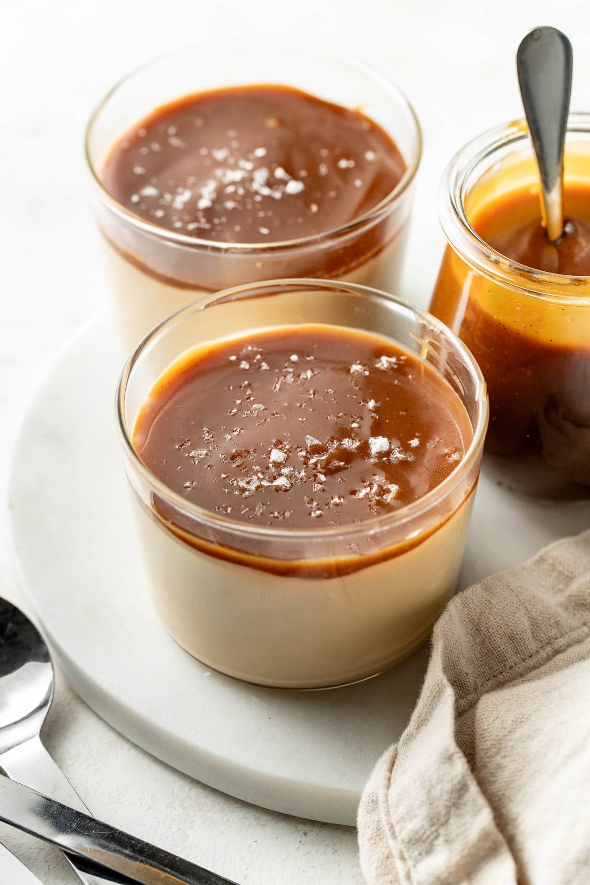 Two glass cups filled with creamy dessert topped with caramel sauce and sprinkled with sea salt, next to a jar of caramel sauce with a spoon, all on a white surface with a beige cloth and spoons nearby.