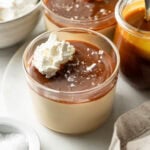 Two glass cups filled with creamy pudding, topped with caramel sauce, flaky sea salt, and whipped cream. A jar of caramel sauce, bowl of whipped cream, and a small dish of sea salt are nearby on a white surface.