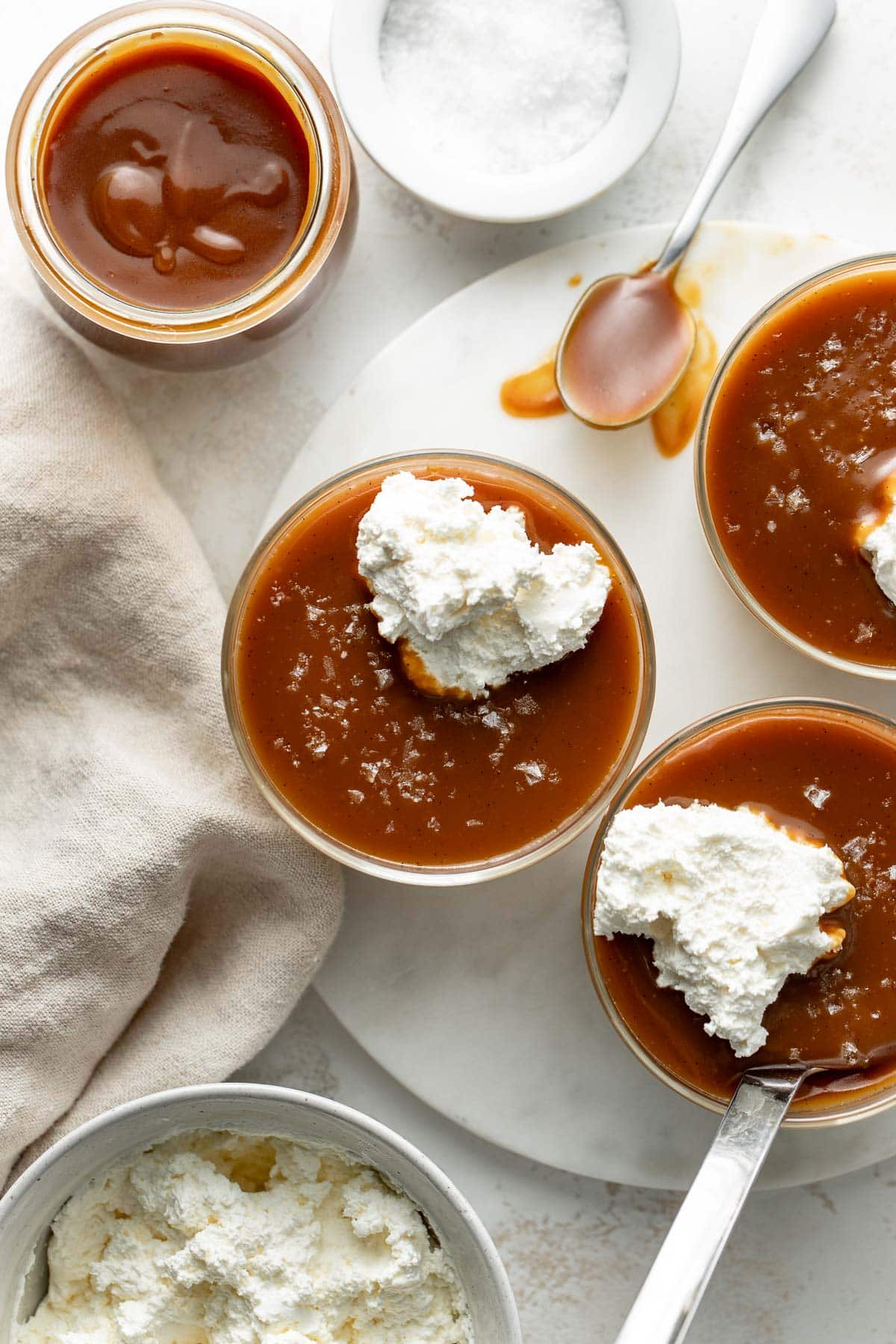 Overhead view of caramel pudding topped with whipped cream and sea salt in glass bowls, with a jar of caramel sauce, a bowl of whipped cream, a small bowl of salt, a spoon, and a beige napkin nearby.