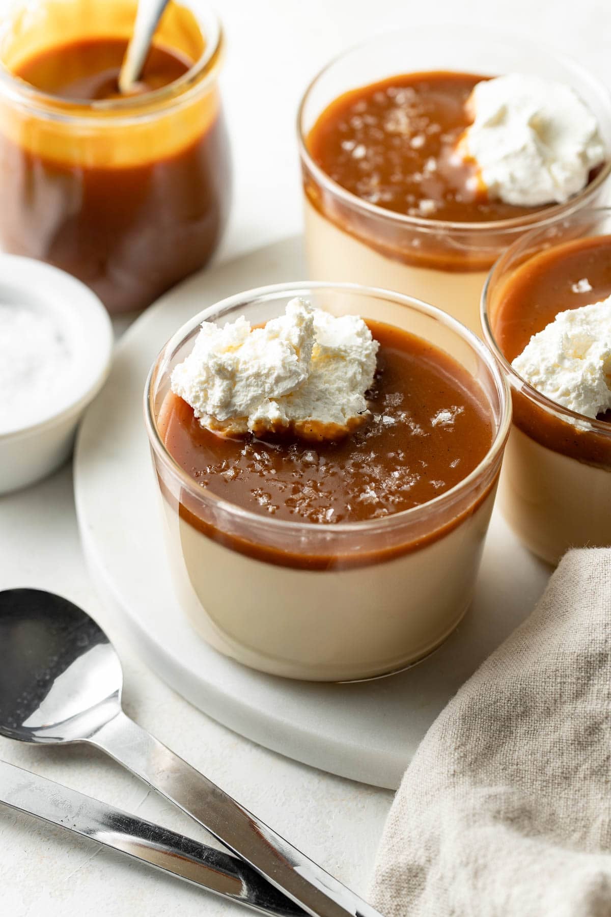 Three glass cups of creamy dessert are topped with caramel sauce, a dollop of whipped cream, and a sprinkle of flaky salt. A jar of caramel sauce, a bowl of salt, spoons, and a beige napkin are nearby.