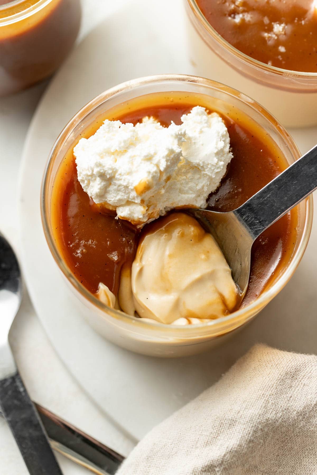 A glass bowl filled with creamy dessert topped with caramel sauce and a dollop of whipped cream. A spoon is scooping some of the dessert, with another bowl and a spoon nearby on a white surface.