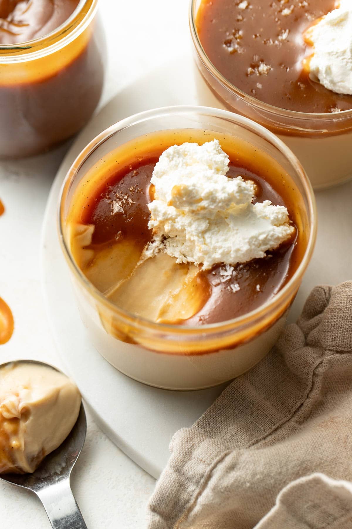 A glass cup filled with creamy pudding topped with caramel sauce, a dollop of whipped cream, and a sprinkle of sea salt sits on a white surface next to a spoon and a beige napkin.
