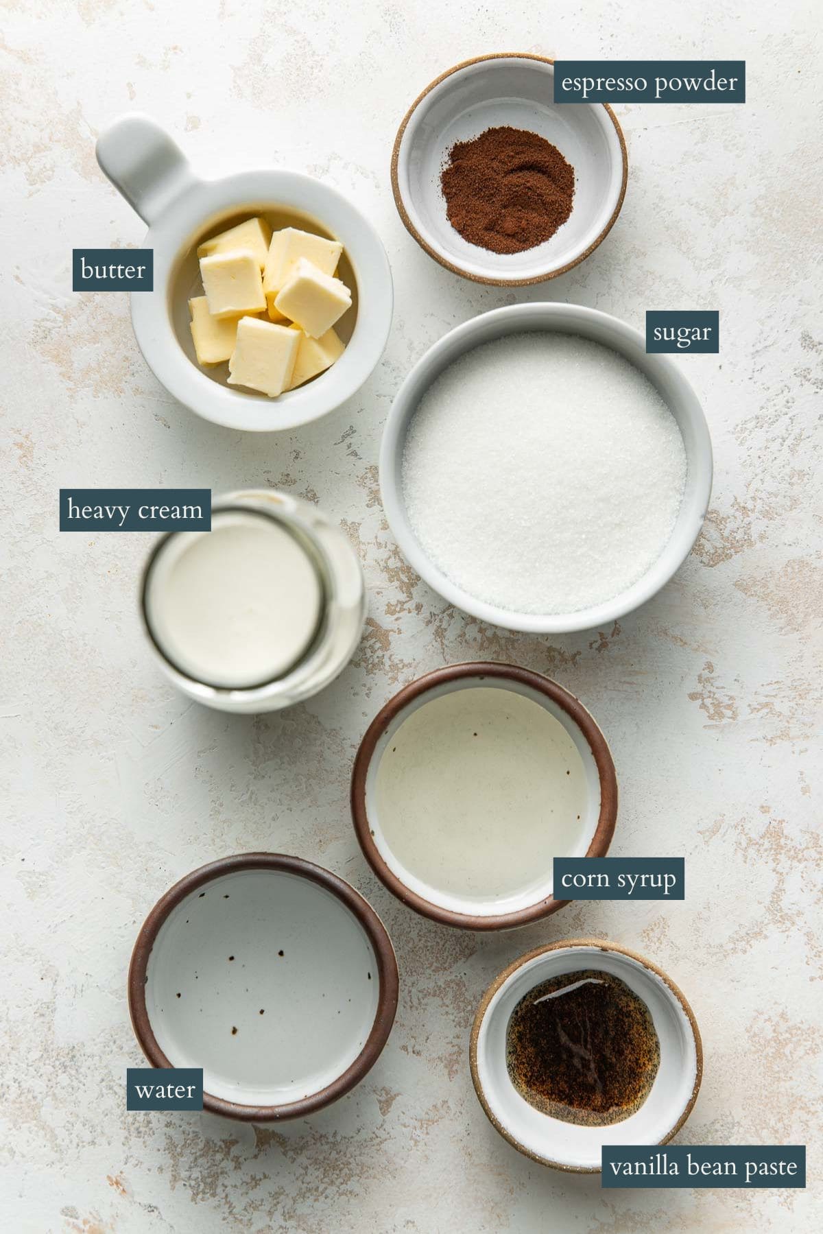 Seven labeled bowls and jars contain ingredients on a light surface: espresso powder, cubed butter, sugar, heavy cream, corn syrup, water, and vanilla bean paste.