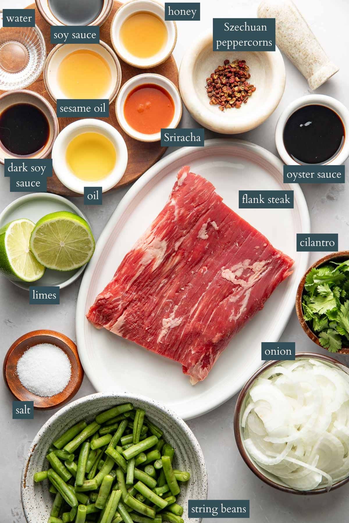 A variety of ingredients for a flank steak dish are arranged on a white surface, including raw flank steak, string beans, sliced onion, cilantro, lime, salt, oil, dark soy sauce, sesame oil, soy sauce, water, honey, Sriracha, oyster sauce, and Szechuan peppercorns.