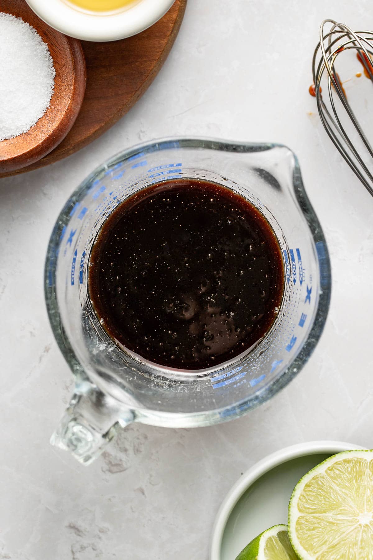 A glass measuring cup filled with a dark liquid sits on a light surface, surrounded by a whisk, a bowl of sliced limes, a bowl of coarse salt, and a partially visible white dish.