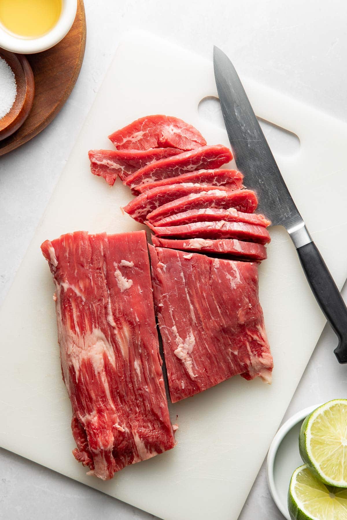 Raw flank steak being sliced on a white cutting board with a chef’s knife. Lime halves and small bowls of oil and salt are nearby on the countertop.