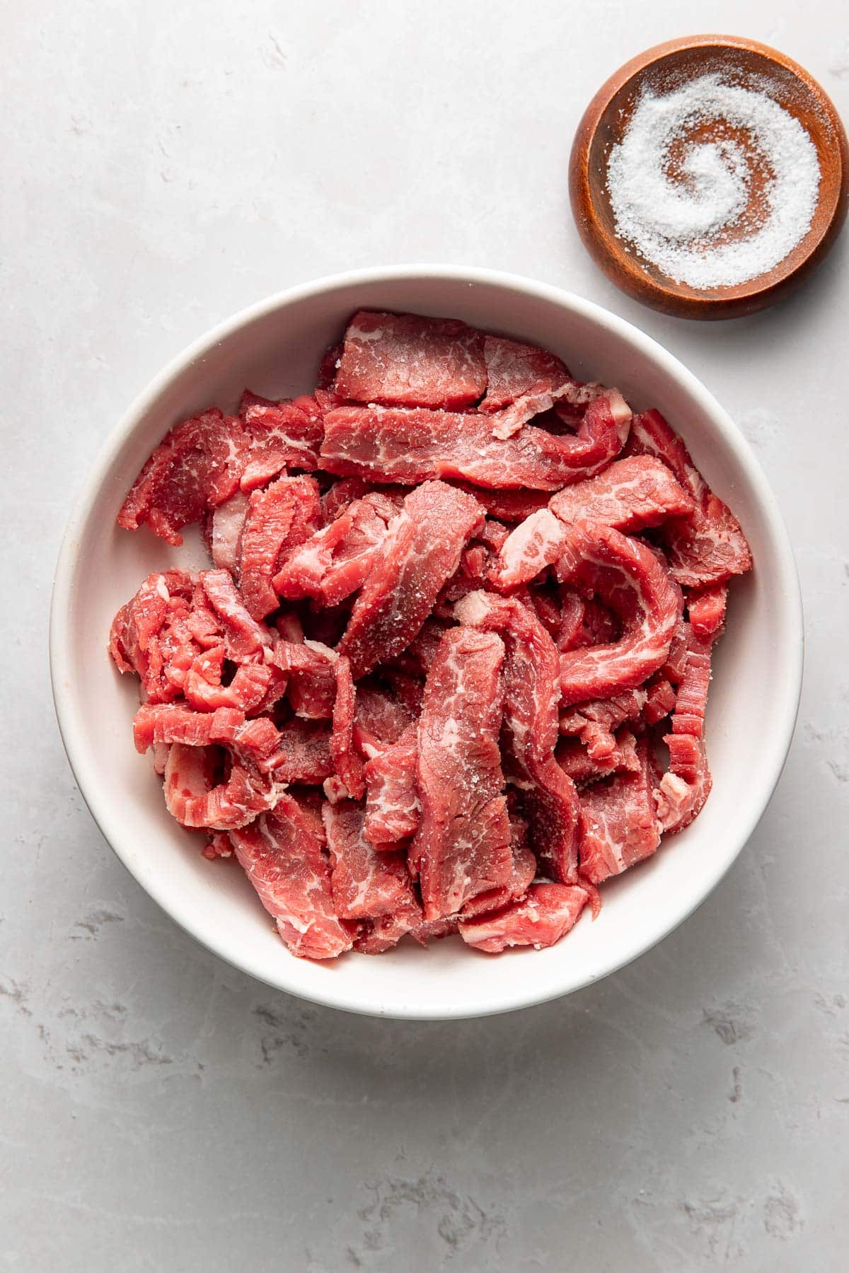 A white bowl filled with raw, sliced beef strips sits on a light surface. Next to it, there is a small wooden bowl containing coarse salt arranged in a spiral pattern.