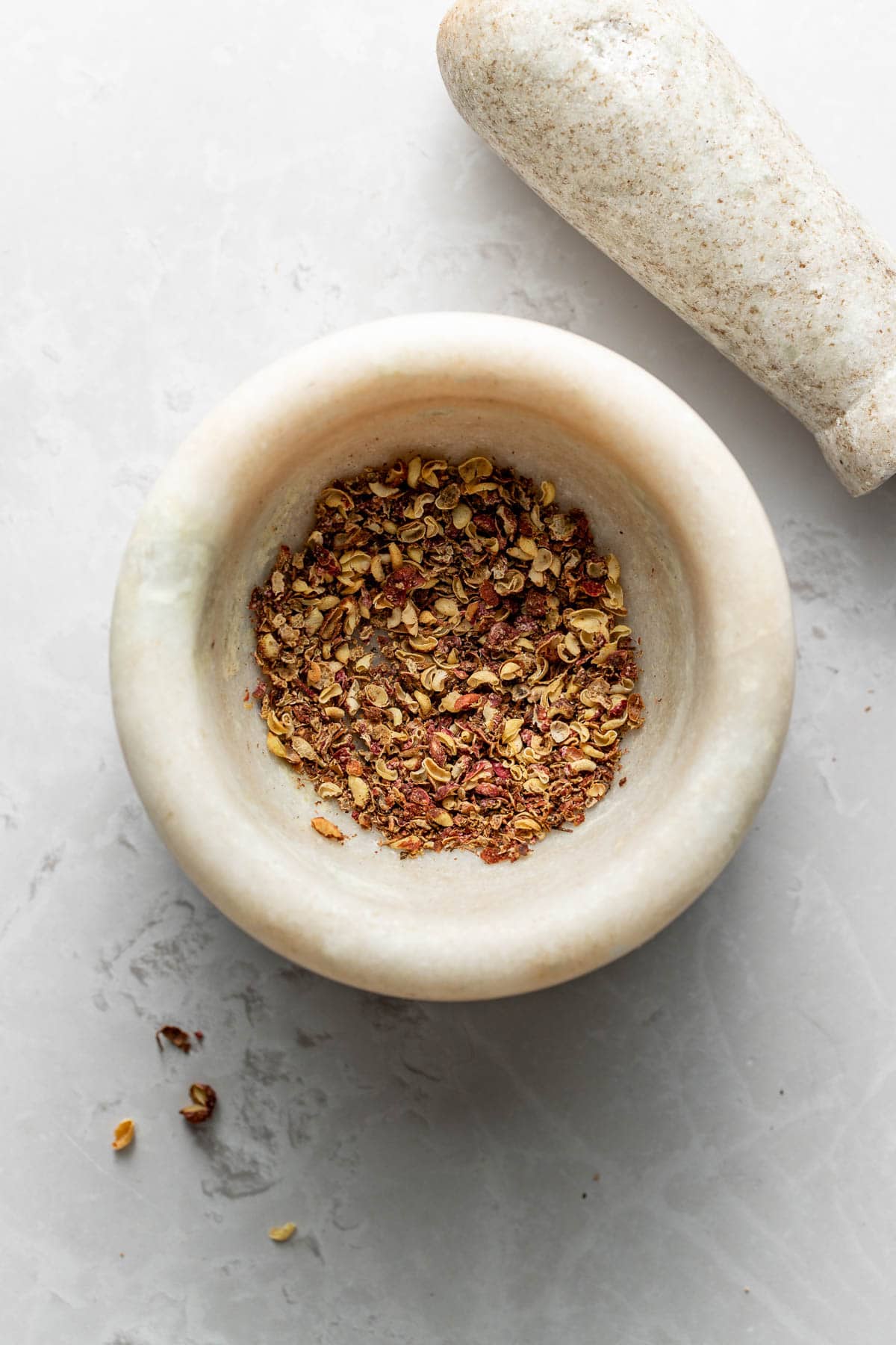 A marble mortar containing crushed spices sits on a light gray surface, with the matching pestle placed nearby. Some spice pieces are scattered beside the mortar.