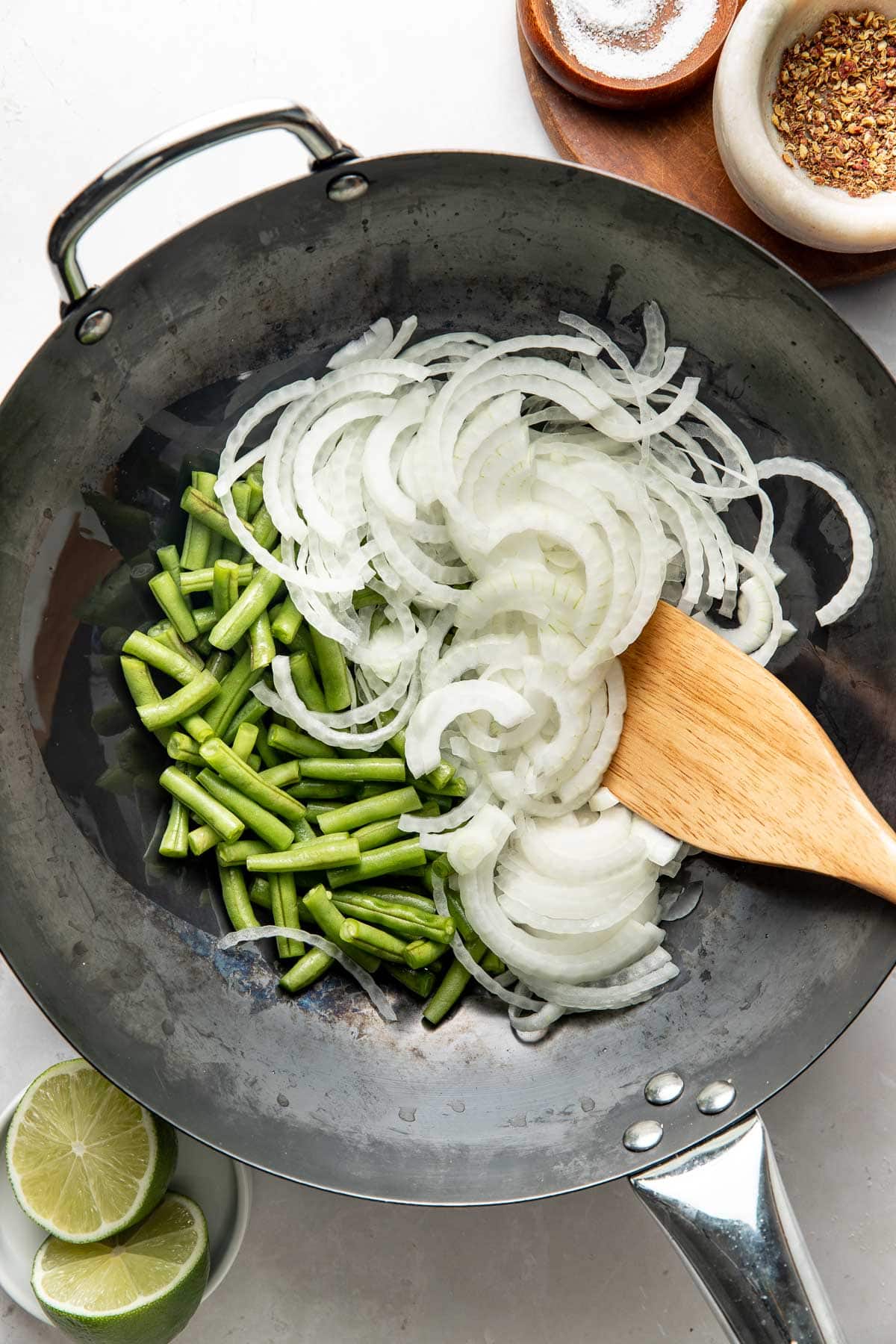 A wok with chopped green beans and sliced onions being stirred with a wooden spatula. Nearby are bowls of salt and red pepper flakes, plus a halved lime on a small plate.