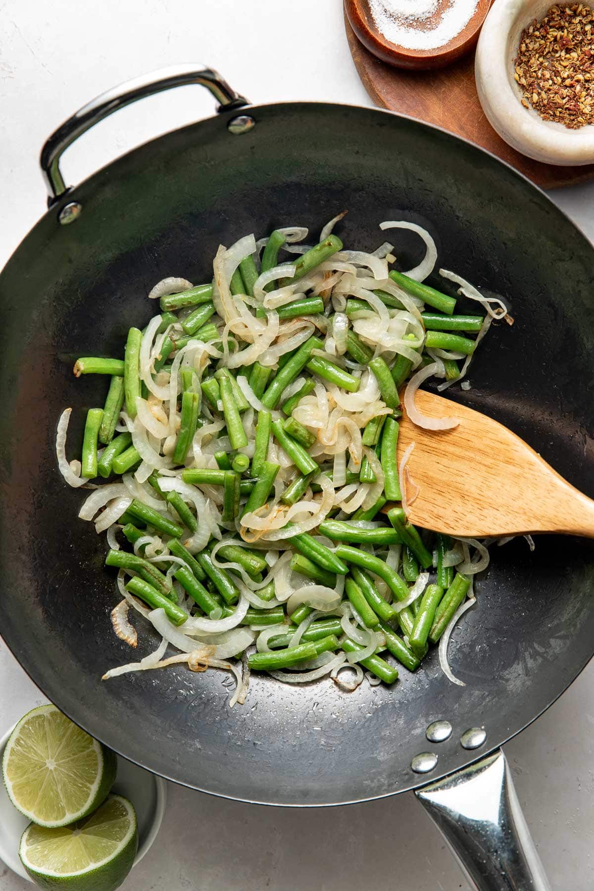 A black wok containing sautéed green beans and sliced onions, with a wooden spatula resting inside. Nearby are lime halves, a bowl of spices, and a small salt dish on a white surface.