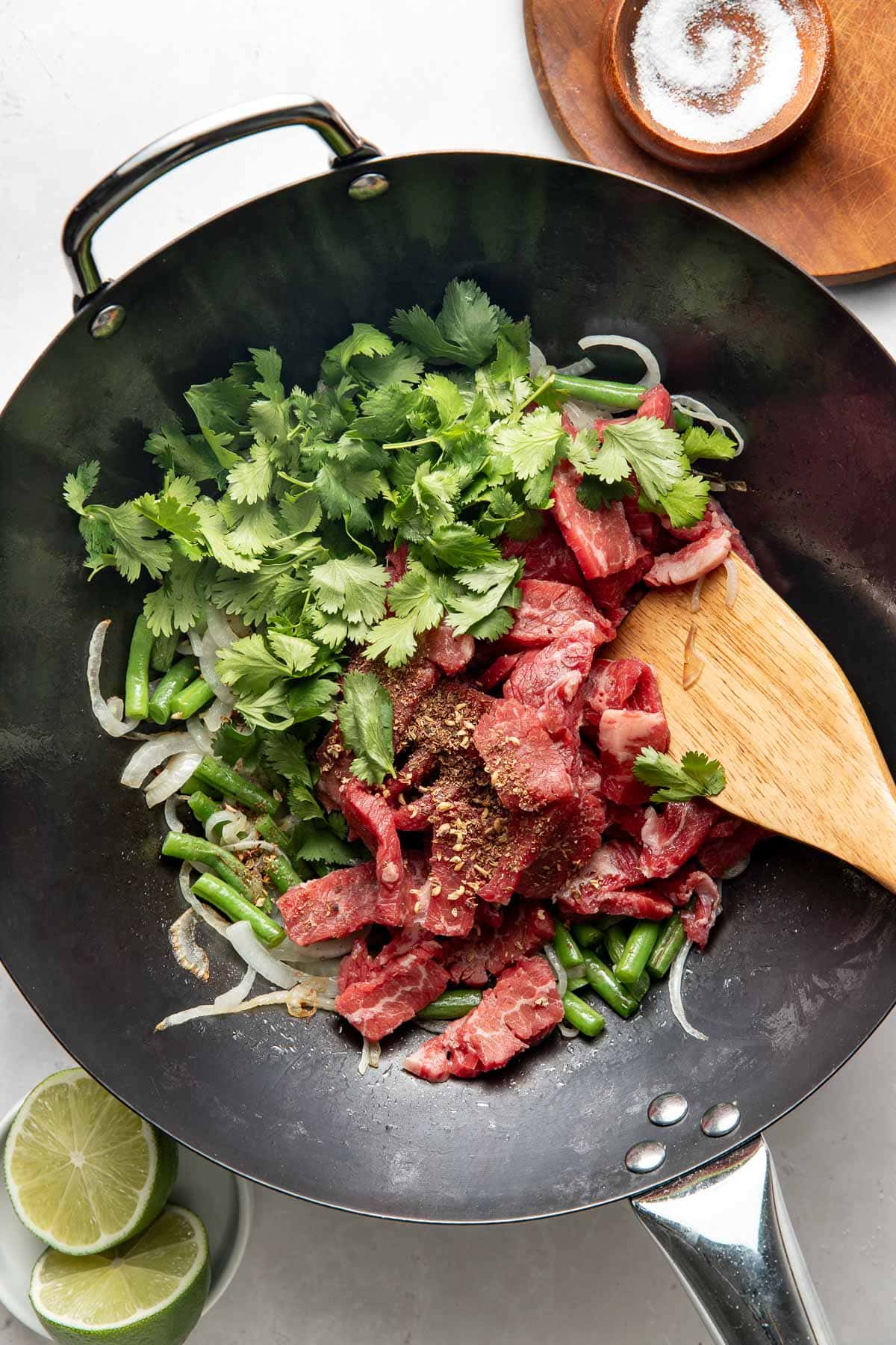 A wok with sliced beef, green beans, onion, fresh cilantro, and spices being stirred with a wooden spatula. Sliced limes and a small bowl of salt are nearby on a white surface.
