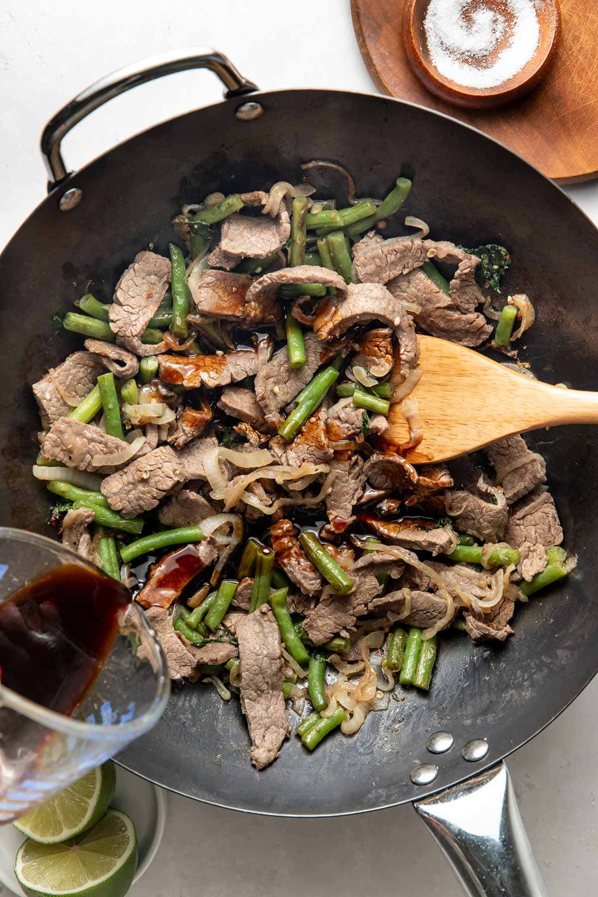 A wooden spoon stirs beef, green beans, and onions in a wok, while a hand pours dark sauce into the pan. A bowl of salt and lime wedges sit nearby on a white surface.