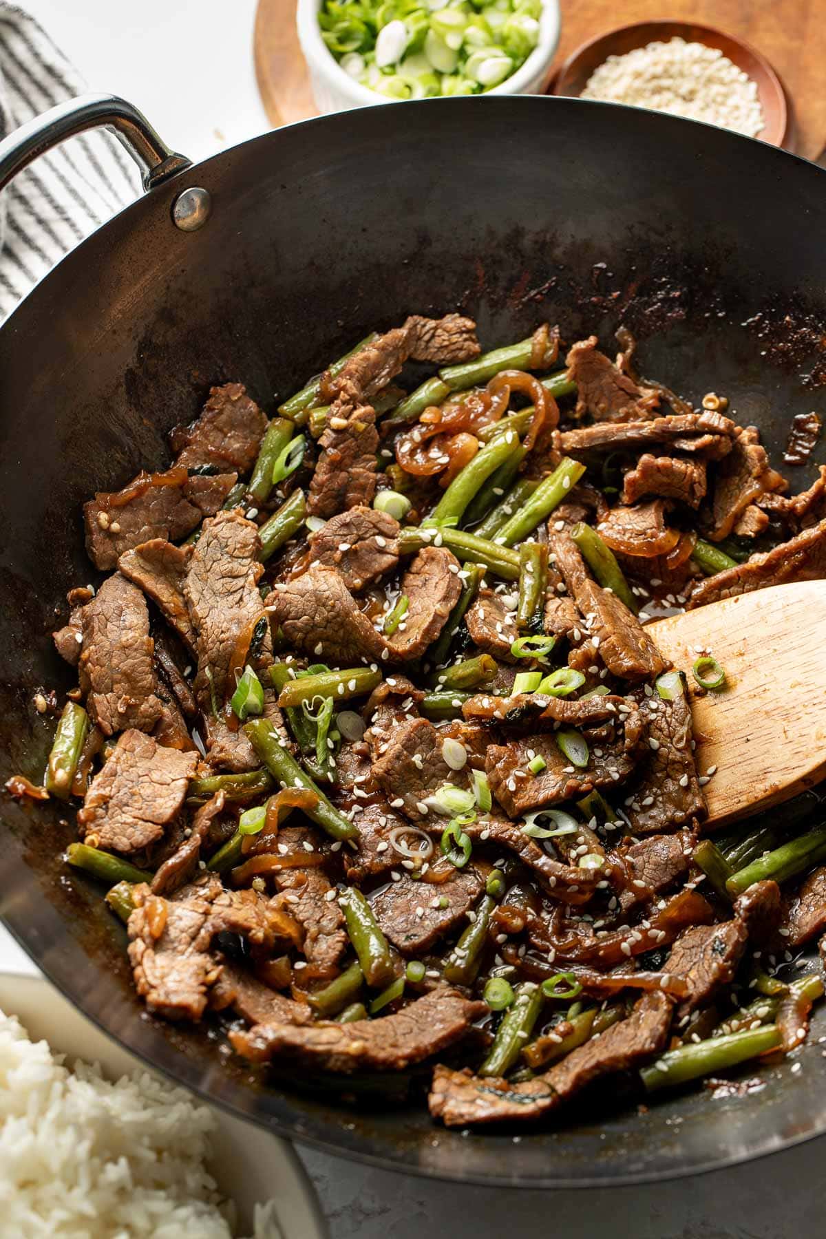 A wok filled with stir-fried beef strips, green beans, and sliced onions, garnished with chopped green onions and sesame seeds, with a bowl of rice and small bowls of sliced green onions and sesame seeds nearby.