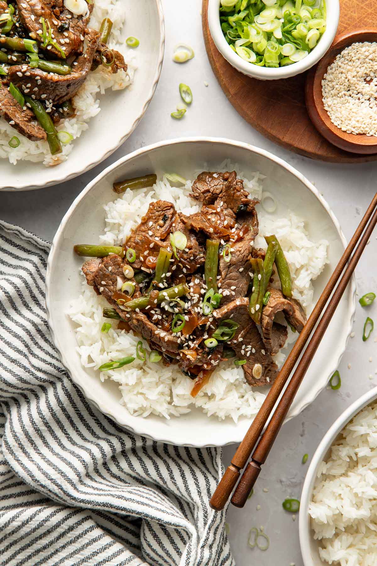 A bowl of white rice topped with sliced beef, green beans, chopped green onions, and sesame seeds, served with chopsticks. A striped cloth napkin, bowls of sliced green onions, and sesame seeds are nearby.