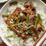 A bowl of white rice topped with sliced beef, green beans, and chopped green onions, garnished with sesame seeds. Chopsticks rest on the bowl, and a small bowl of green onions is nearby.