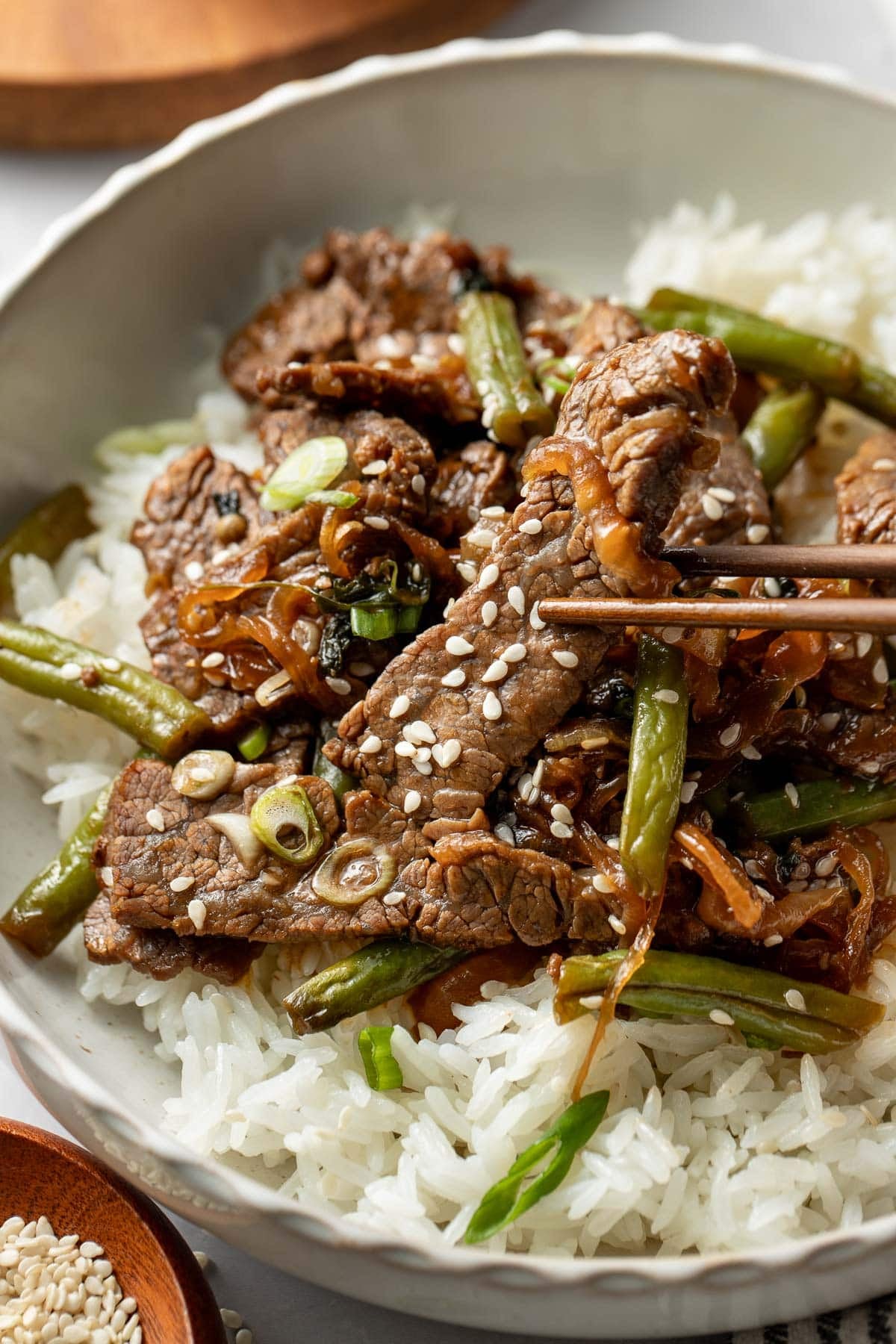 A bowl of white rice topped with stir-fried beef, green beans, onions, and sesame seeds, with chopsticks picking up a piece of beef.