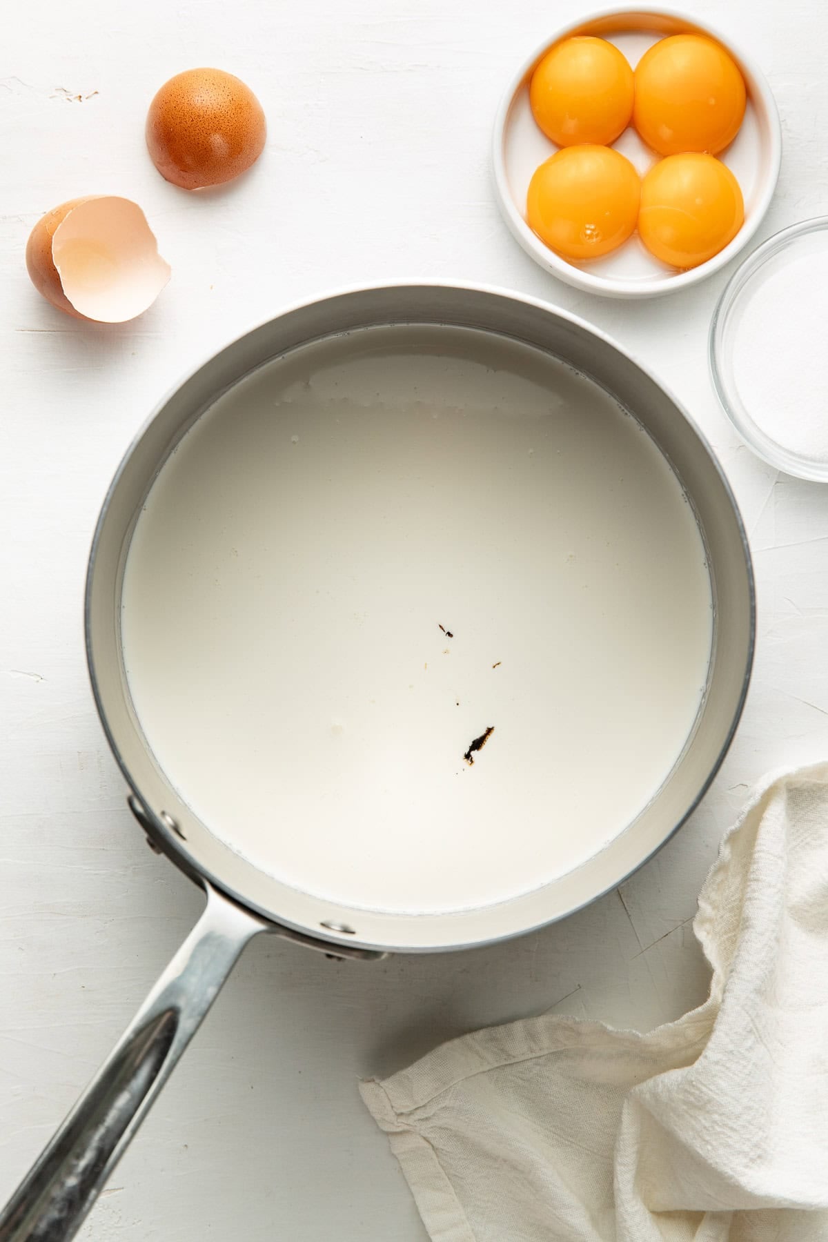 A saucepan filled with milk, next to an empty eggshell, three egg yolks on a small plate, a bowl of sugar, and a white cloth on a white surface.